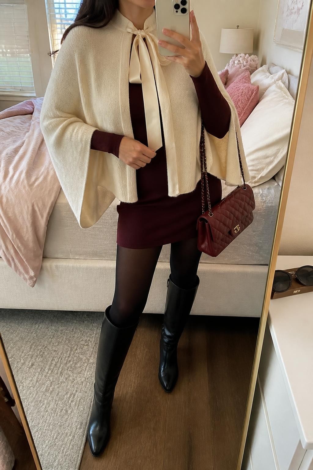 Image Prompt mirror selfie of a woman wearing a cream soft-knit cape cardigan with a loosely tied satin neck ribbon over a burgundy fitted long-sleeve mini dress, black opaque tights, black leather knee-high boots, and a burgundy quilted shoulder bag, clearly showing the exact clothing items, colors, fabrics, ribbon, tights, boots, and bag listed in the Outfit Pieces section, holding a phone in front of a mirror, cropped at NECK so the face does not show, relaxed feminine selfie pose in a feminine-themed bedroom, soft indoor lighting, with soft pillows on an unmade bed and sunglasses placed on a dresser, realistic soft knit, satin, dress fabric, tights, leather, and quilting textures, latest iPhone photo quality