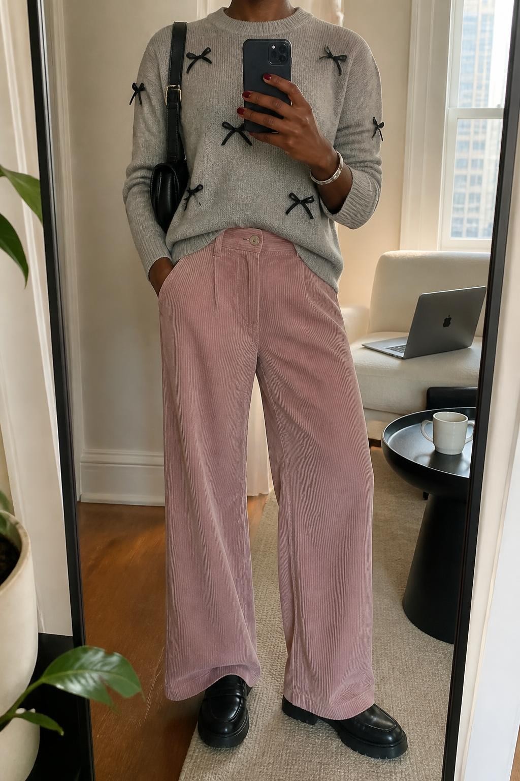 Image Prompt mirror selfie of a Black woman wearing a soft gray crewneck sweater with small black satin bows and a small front tuck into dusty blush wide-leg corduroy trousers, black chunky leather loafers, a black structured shoulder bag, and a slim silver cuff bracelet, clearly showing the exact clothing items, colors, fabrics, bows, shoes, bag, and bracelet listed in the Outfit Pieces section, holding a phone in front of a mirror, cropped at NECK so the face does not show, relaxed casual selfie pose in a stylish city apartment, natural window light, with a coffee mug on a side table and a laptop resting on a chair, realistic knit, satin, corduroy, leather, and silver textures, latest iPhone photo quality