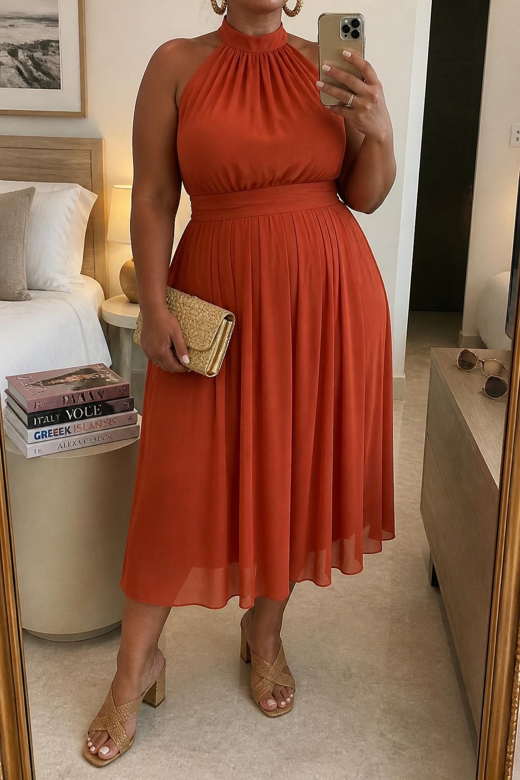 Mirror selfie of a plus size woman wearing a burnt orange chiffon halter-neck midi dress with a wide supportive neck band, a fitted waist, and a softly pleated skirt, tan woven block-heel sandals, a natural raffia clutch with gold trim, and hammered gold hoop earrings, clearly showing the exact dress shape, burnt orange color, chiffon texture, footwear, and accessories listed, holding a phone in front of a mirror, cropped at the neck so her face does not show, relaxed graceful selfie pose, in a cozy bedroom mirror setup with soft indoor lighting, a stack of fashion magazines on a side table and sunglasses placed on a dresser, realistic destination wedding appearance, latest iPhone photo quality.