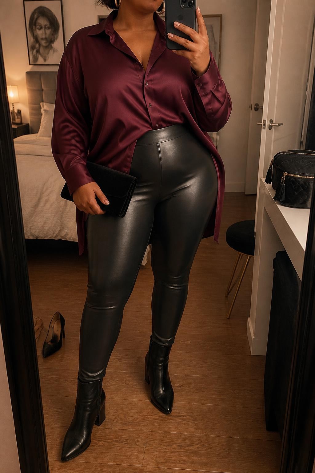 Mirror selfie of a plus size woman wearing an oversized burgundy satin button-down shirt with a long curved hem, black high-waisted faux-leather leggings, black pointed ankle boots with block heels, a small black velvet clutch, and chunky gold hoop earrings, clearly showing the exact clothing items, burgundy satin, black leather texture, footwear, and accessories listed, holding a phone in front of a mirror, cropped at the neck so her face does not show, relaxed bold selfie stance, in a cozy bedroom mirror setup with warm moody lighting, a pair of heels on the floor nearby and a cosmetic bag on the counter, realistic date-night outfit, latest iPhone photo quality.
