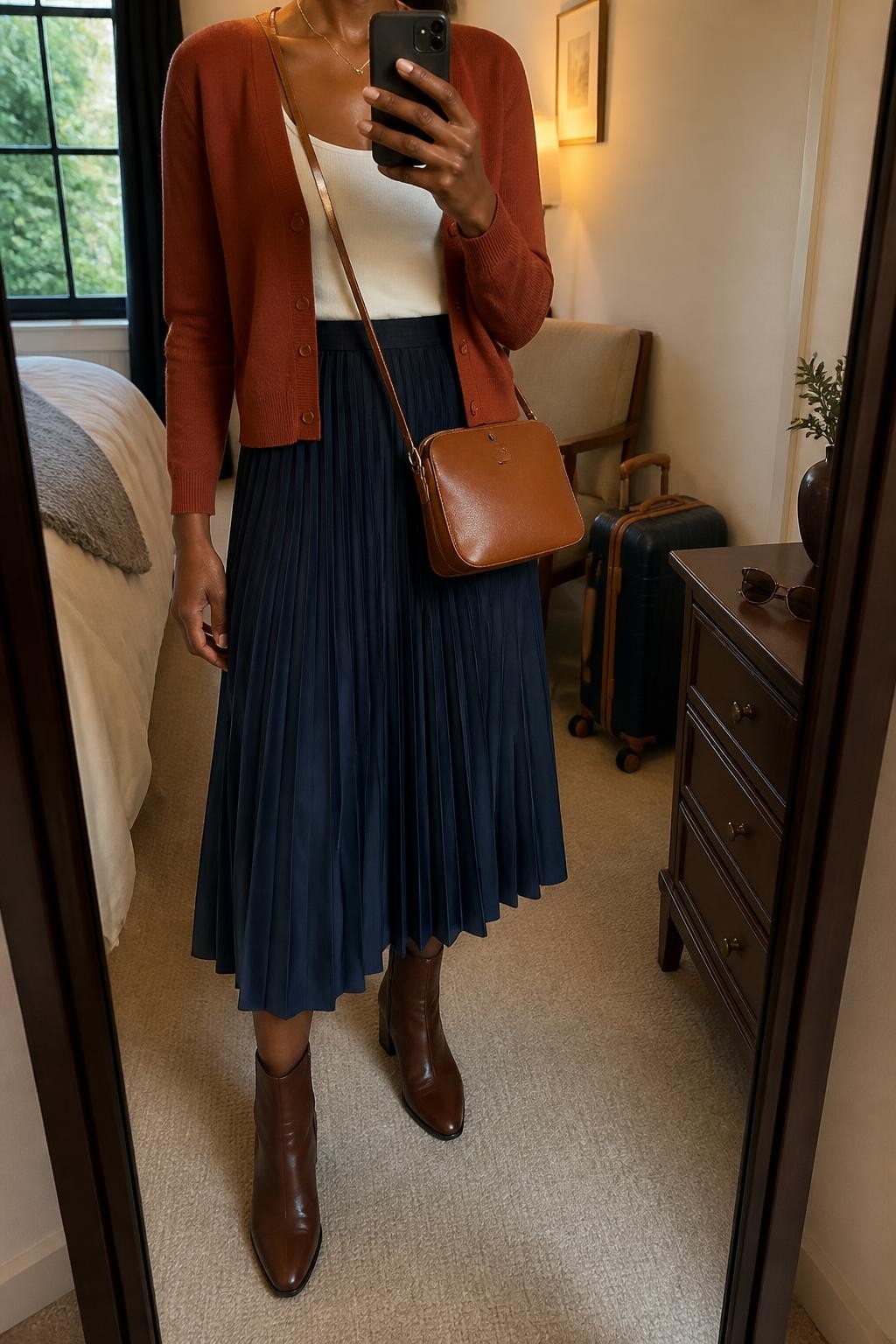 mirror selfie of a Black woman wearing a rust fine-knit V-neck cardigan buttoned halfway over an ivory fitted scoop-neck top tucked into a navy pleated midi skirt, chocolate leather ankle boots, and a cognac zip-top crossbody bag, clearly showing the exact clothing items, colors, fabrics, boots, and bag listed in the Outfit Pieces section, holding a phone in front of a mirror, cropped at NECK so the face does not show, relaxed feminine selfie pose in a cozy bedroom mirror setup, evening golden hour light, with a small suitcase beside a chair and sunglasses placed on a dresser, realistic fine knit, pleated fabric, and leather textures, latest iPhone photo quality