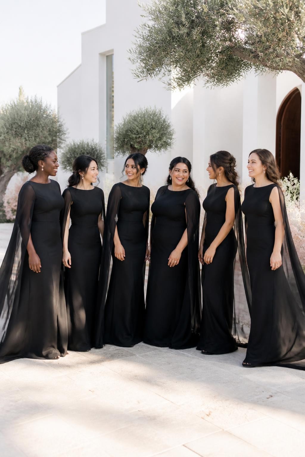 Realistic editorial wedding photo of six bridesmaids in black chiffon gowns with clean fitted bodices and sheer cape sleeves flowing from the shoulders to the floor, women of West African, East Asian, South Asian, Middle Eastern, Latina, and European appearance with varied skin tones, heights, ages, and body types, bright expressive faces, subtle natural makeup, soft flyaway hairs and individual facial features, standing beside a modern white wedding chapel surrounded by olive trees, cream stone flooring, pale peach florals, and soft afternoon sunshine, light breeze lifting the chiffon capes naturally, realistic shadows, delicate fabric transparency, no stiff posing, high-detail DSLR photography.