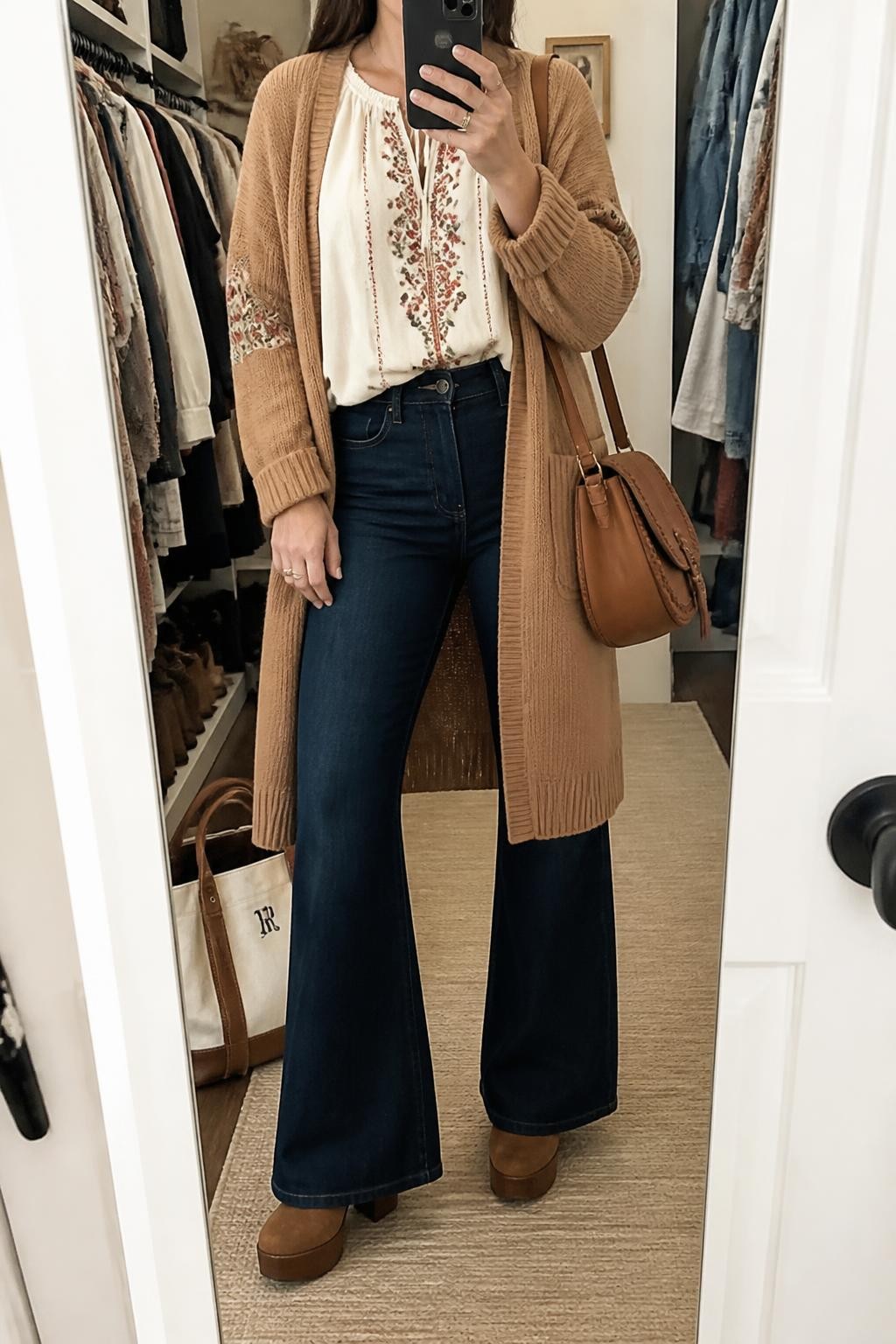 Mirror selfie of a woman wearing dark-wash high-waisted flared jeans, an ivory cotton peasant blouse with rust and olive embroidery, a long camel knit cardigan, brown suede platform ankle boots, and a cognac leather shoulder bag, clearly showing the exact clothing items, colors, fabrics, and accessories listed, holding a phone in front of a walk-in closet mirror, cropped at the neck so the face does not show, relaxed casual selfie pose, soft indoor lighting, an open closet with clothes visible and a tote bag resting on the floor, natural everyday setting, latest iPhone photo quality.