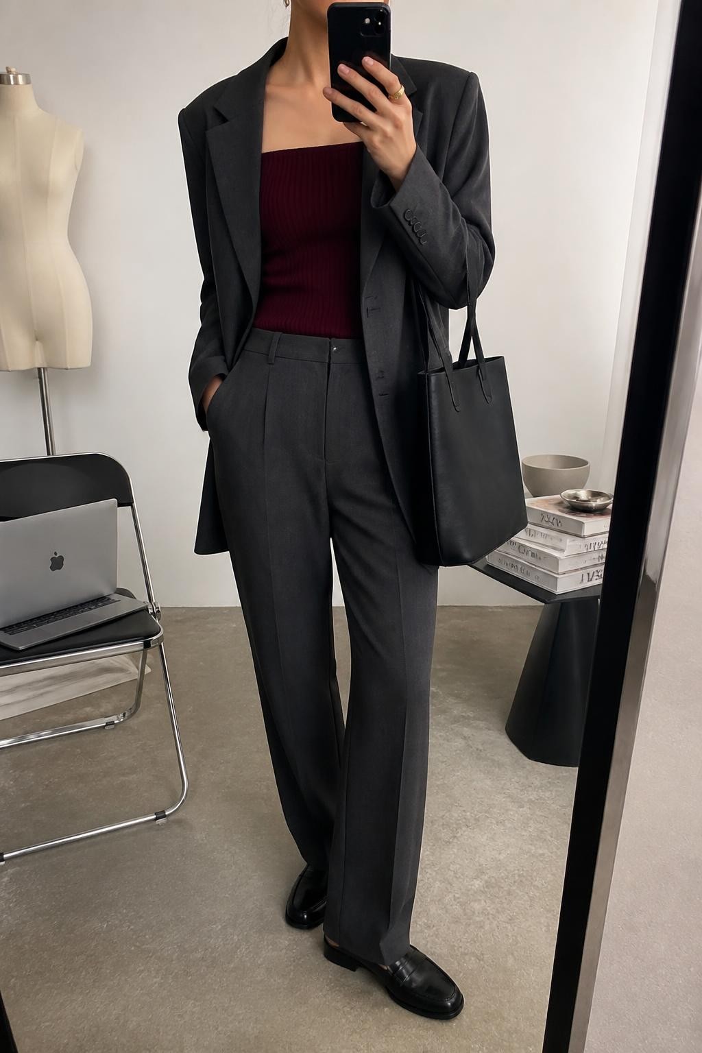 mirror selfie of a woman wearing an open charcoal relaxed-fit tailored blazer over a burgundy fitted square-neck knit top tucked into charcoal straight-leg tailored trousers, black polished leather loafers, and a black structured work tote, clearly showing the exact clothing items, colors, fabrics, shoes, and bag listed in the Outfit Pieces section, holding a phone in front of a mirror, cropped at NECK so the face does not show, relaxed authoritative selfie pose in a fashion studio mirror corner, soft indoor lighting, with a stack of fashion magazines on a side table and a laptop on a chair, realistic tailored, knit, and polished leather textures, latest iPhone photo quality