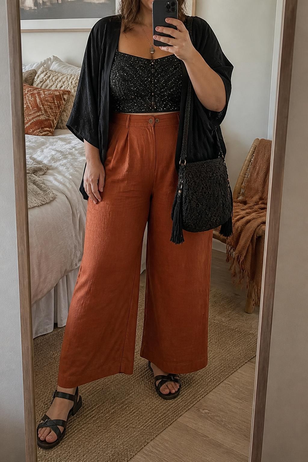A realistic fashion mirror selfie of a plus-size adult woman wearing a warm-weather festival outfit: a full-coverage black beaded cropped top, rust-orange high-waisted wide-leg linen pants, and a lightweight black cropped kimono layered over the outfit. She wears flat black leather sandals with secure ankle straps and carries a black woven shoulder bag. She is holding a smartphone in front of a full-length mirror, with the composition cropped below the chin so her face is not visible. Relaxed, casual fashion pose in a cozy bedroom mirror setup with soft indoor lighting, soft pillows on an unmade bed and a folded blanket on a chair. Realistic contemporary festival styling, detailed beadwork and linen texture, warm earthy colors, natural proportions, and high-quality modern smartphone photography.