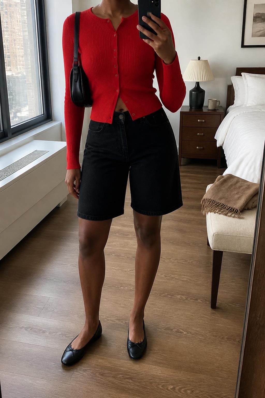 mirror selfie of a Black woman wearing high-waisted structured black denim jorts, a fitted red fine-knit cardigan with small buttons and the bottom button left open slightly, rounded-toe black ballet flats, a smooth black leather mini shoulder bag and small thick gold hoop earrings, wearing exactly the listed outfit with no added garments, holding a phone in front of a mirror, cropped at NECK so the face does not show, relaxed casual selfie pose, in a stylish city apartment, natural window light, coffee mug on a bedside table and a folded blanket on a chair, realistic unposed latest iPhone photo quality.