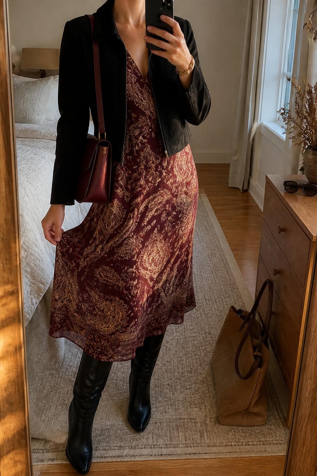 mirror selfie of a woman wearing a burgundy and gold paisley-print midi dress, an open black cropped suede jacket, black leather knee-high boots, a burgundy structured shoulder bag, and an antique gold bracelet, clearly showing the exact clothing items, colors, paisley print, fabrics, boots, bag, and bracelet listed in the Outfit Pieces section, holding a phone in front of a mirror, cropped at NECK so the face does not show, relaxed feminine selfie pose in a cozy bedroom mirror setup, evening golden hour light, with a tote bag resting on the floor and sunglasses placed on a dresser, realistic printed dress fabric, suede, leather, and metal textures, latest iPhone photo quality