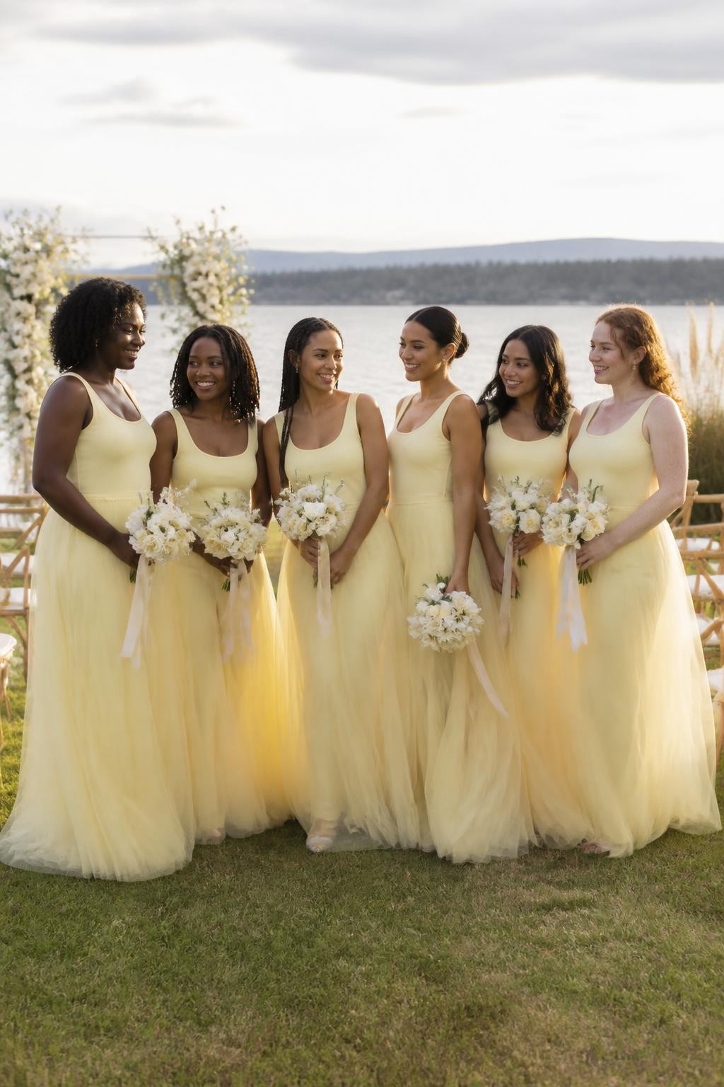 Realistic romantic lakeside wedding photograph of six bridesmaids wearing pale limoncello yellow tulle gowns with clean fitted scoop-neck bodices, elongated dropped waists, and airy layered skirts beginning around the upper hips, women with deep ebony, rich warm brown, medium brown, golden tan, South Asian olive, and fair freckled skin tones, varied torso lengths, heights, ages, and body shapes represented naturally, bright expressive faces with realistic skin texture, hairstyles including natural curls, shoulder-length locs, long braids, smooth low bun, layered waves, and short coils, standing beside a lakeside ceremony with cream flowers, pale ribbons, wooden chairs, tall grasses, and calm water, gentle wind moving the tulle naturally, soft golden-hour light passing through the layers, visible fabric edges, believable transparency, and accurate anatomy.