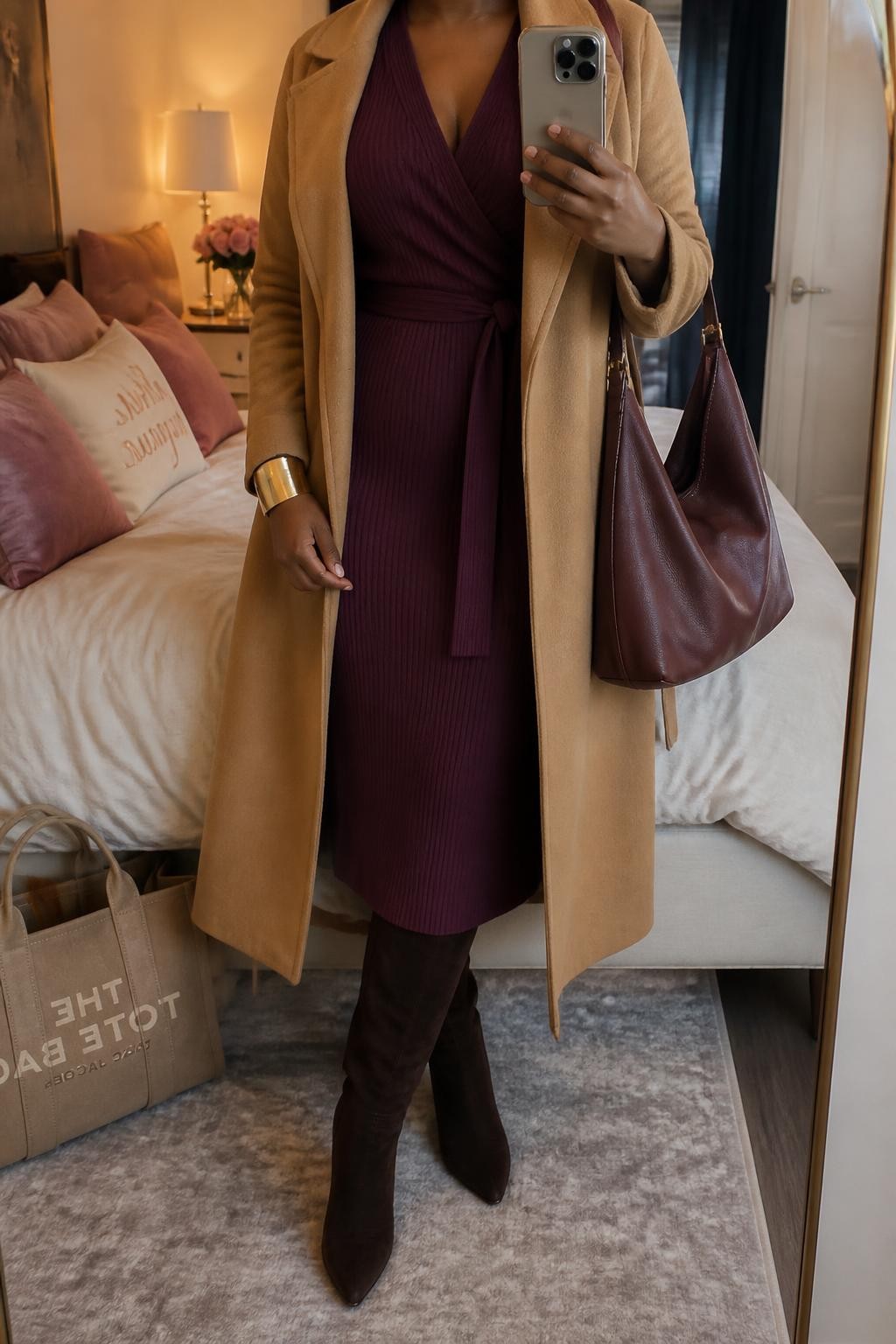 mirror selfie of a Black midsize woman wearing a deep plum ribbed knit wrap midi dress tied securely at the natural waist, an open camel belted wool-blend coat, chocolate suede knee-high boots, an oxblood leather shoulder bag, and a wide gold cuff bracelet, clearly showing the exact clothing items, colors, fabrics, boots, bag, and bracelet listed in the Outfit Pieces section, holding a phone in front of a mirror, cropped at NECK so the face does not show, relaxed polished selfie pose in a feminine-themed bedroom, evening golden hour light, with a tote bag resting on the floor and soft pillows on an unmade bed, realistic ribbed knit, wool, suede, and leather textures, natural stance and mirror reflection, latest iPhone photo quality