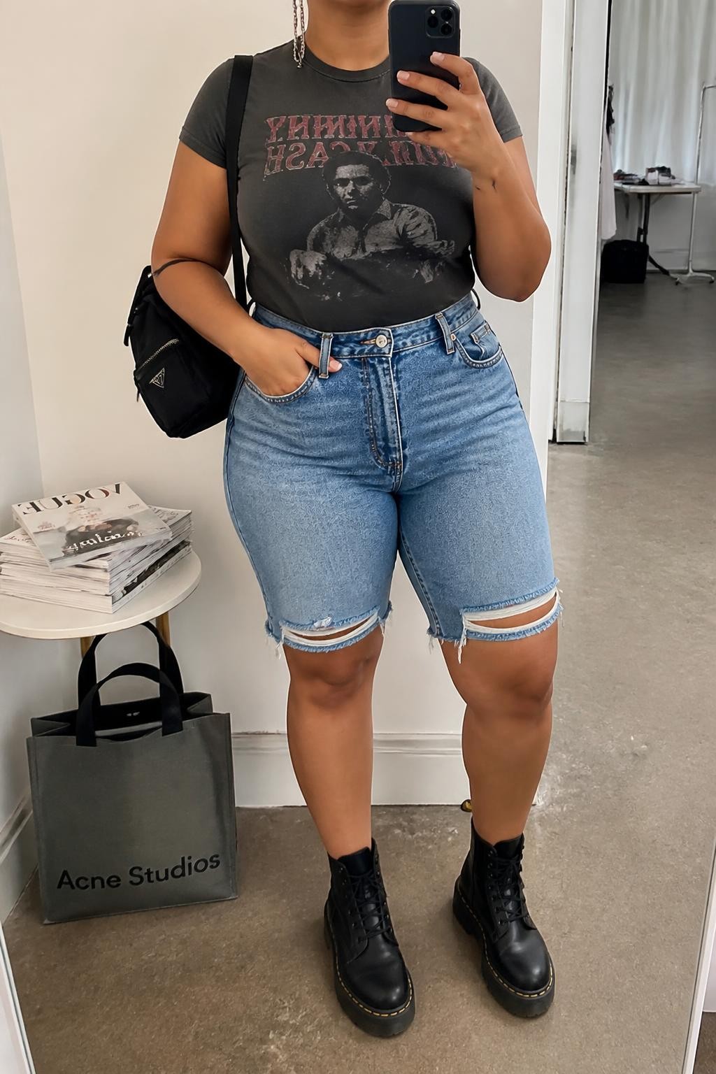 Mirror selfie of a plus size woman wearing medium-wash high-waisted distressed denim jorts with small frayed knee details, a fitted faded charcoal graphic T-shirt tucked into the waistband, black lace-up combat boots, a compact black nylon backpack, and silver chain earrings, clearly showing the exact clothing items, denim distressing, graphic print, black footwear, and accessories listed, holding a phone in front of a mirror, cropped at the neck so her face does not show, relaxed edgy selfie stance, in a fashion studio mirror corner with soft indoor lighting, a stack of fashion magazines on a side table and a tote bag resting on the floor, realistic festival outfit, latest iPhone photo quality.