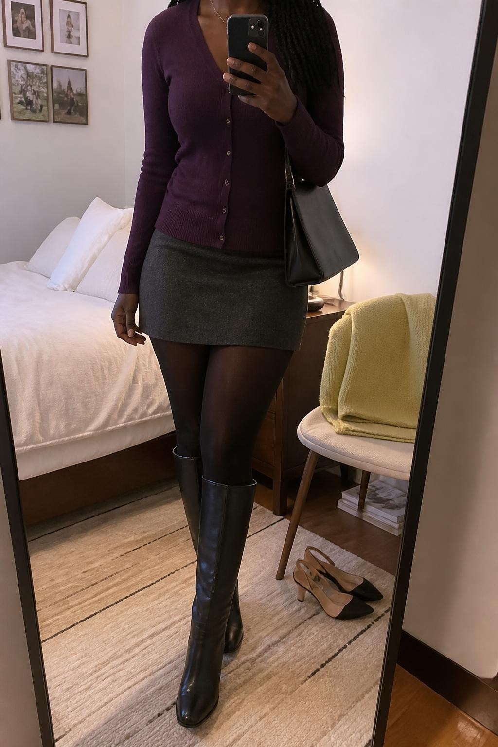 Mirror selfie of a Black woman wearing a fitted plum-purple cardigan, a charcoal-grey wool mini skirt, opaque black tights, black leather knee-high boots, and a structured black shoulder bag, clearly showing the exact clothing items, colors, fabrics, and accessories listed, holding a phone in front of a cozy bedroom mirror setup, cropped at the neck so the face does not show, relaxed casual selfie pose, soft indoor lighting, a folded blanket on a chair and a pair of heels on the floor nearby, natural lived-in atmosphere, latest iPhone photo quality.