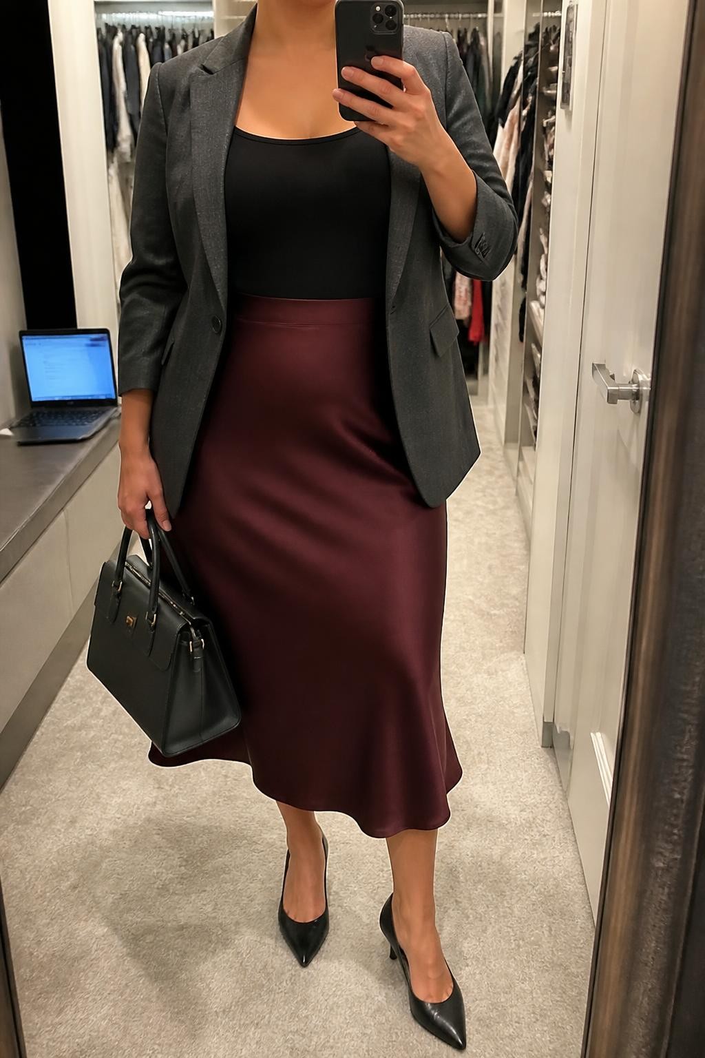 Mirror selfie of a plus size woman wearing a burgundy high-waisted satin midi skirt with a softly flared shape, a fitted black scoop-neck camisole with wide straps, a charcoal grey tailored blazer worn open, black pointed kitten heels, and a structured charcoal handbag, clearly showing the exact clothing items, burgundy satin, black camisole, charcoal blazer, footwear, and accessories listed, holding a phone in front of a mirror, cropped at the neck so her face does not show, relaxed professional selfie stance, in a walk-in closet mirror with soft indoor lighting, a laptop resting on a bench and an open closet with clothes visible, realistic office event outfit, latest iPhone photo quality.