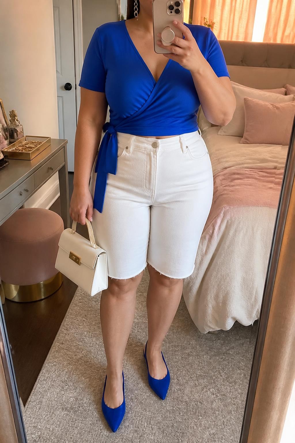 Mirror selfie of a plus size woman wearing crisp white high-waisted denim jorts with a tailored knee-skimming cut, a cobalt blue short-sleeve wrap top with a softly tied waist, cobalt pointed slingback flats, a structured white mini handbag, and silver drop earrings, clearly showing the exact clothing items, white denim, cobalt fabric, wrap shape, colors, footwear, and accessories listed, holding a phone in front of a mirror, cropped at the neck so the face does not appear, relaxed bright selfie pose, in a feminine-themed bedroom with evening golden-hour lighting, soft pillows on an unmade bed and a jewelry tray on the vanity, realistic summer lunch outfit, latest iPhone photo quality.