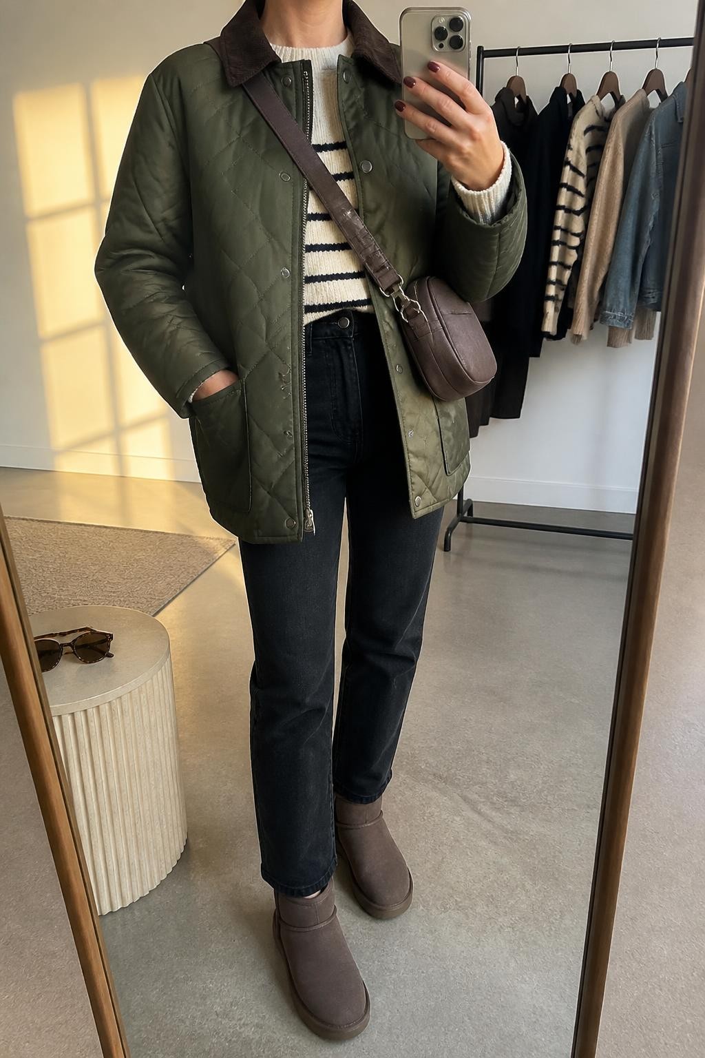 mirror selfie of a woman wearing an olive diamond-quilted barn jacket partly open over a cream and navy striped wool-blend sweater, black high-rise straight-leg jeans tucked neatly into dark brown weatherproof Ugg ankle boots, and a chocolate water-resistant crossbody bag, clearly showing the exact clothing items, colors, pattern, fabrics, boots, and bag listed in the Outfit Pieces section, holding a phone in front of a mirror, cropped at NECK so the face does not show, relaxed outdoor-ready selfie pose in a fashion studio mirror corner, evening golden hour light, with a clothing rack holding a few garments behind her and sunglasses placed on a side table, realistic quilting, wool knit, denim, weatherproof suede, and bag textures, latest iPhone photo quality