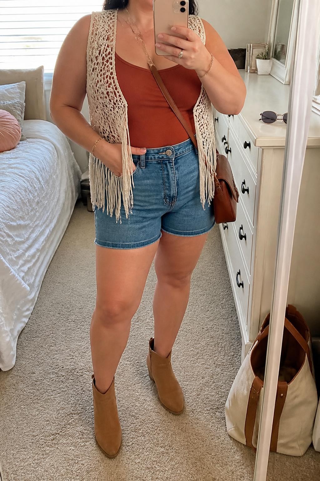 Mirror selfie of a plus size woman wearing a cream crochet fringe top layered over a fitted rust-colored camisole, high-waisted medium-wash denim shorts with a comfortable mid-thigh length, tan suede ankle boots, and a small rust leather crossbody bag, clearly showing the exact clothing items, colors, fabrics, fringe details, footwear, and accessories listed, holding a phone in front of a mirror, cropped at the neck so the face does not show, relaxed casual selfie pose, in a feminine-themed bedroom with natural window light, sunglasses placed on a dresser and a tote bag resting on the floor, natural unstaged festival appearance, latest iPhone photo quality.