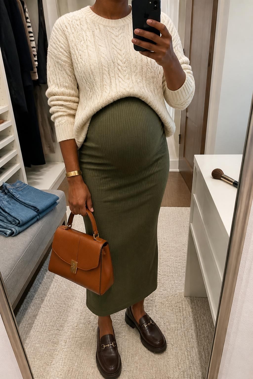 mirror selfie of a Black pregnant woman wearing a cream lightweight cable-knit sweater with a loose front tuck above her visible baby bump, an olive ribbed over-the-bump maternity midi skirt, chocolate cushioned leather loafers, a cognac top-handle bag, and a slim gold cuff bracelet, clearly showing the exact clothing items, colors, fabrics, shoes, bag, and bracelet listed in the Outfit Pieces section, holding a phone in front of a mirror, cropped at NECK so the face does not show, relaxed polished maternity selfie pose in a walk-in closet mirror, soft indoor lighting, with folded denim jeans on a bench and a makeup brush on the vanity, realistic cable knit, ribbed stretch fabric, and leather textures, latest iPhone photo quality