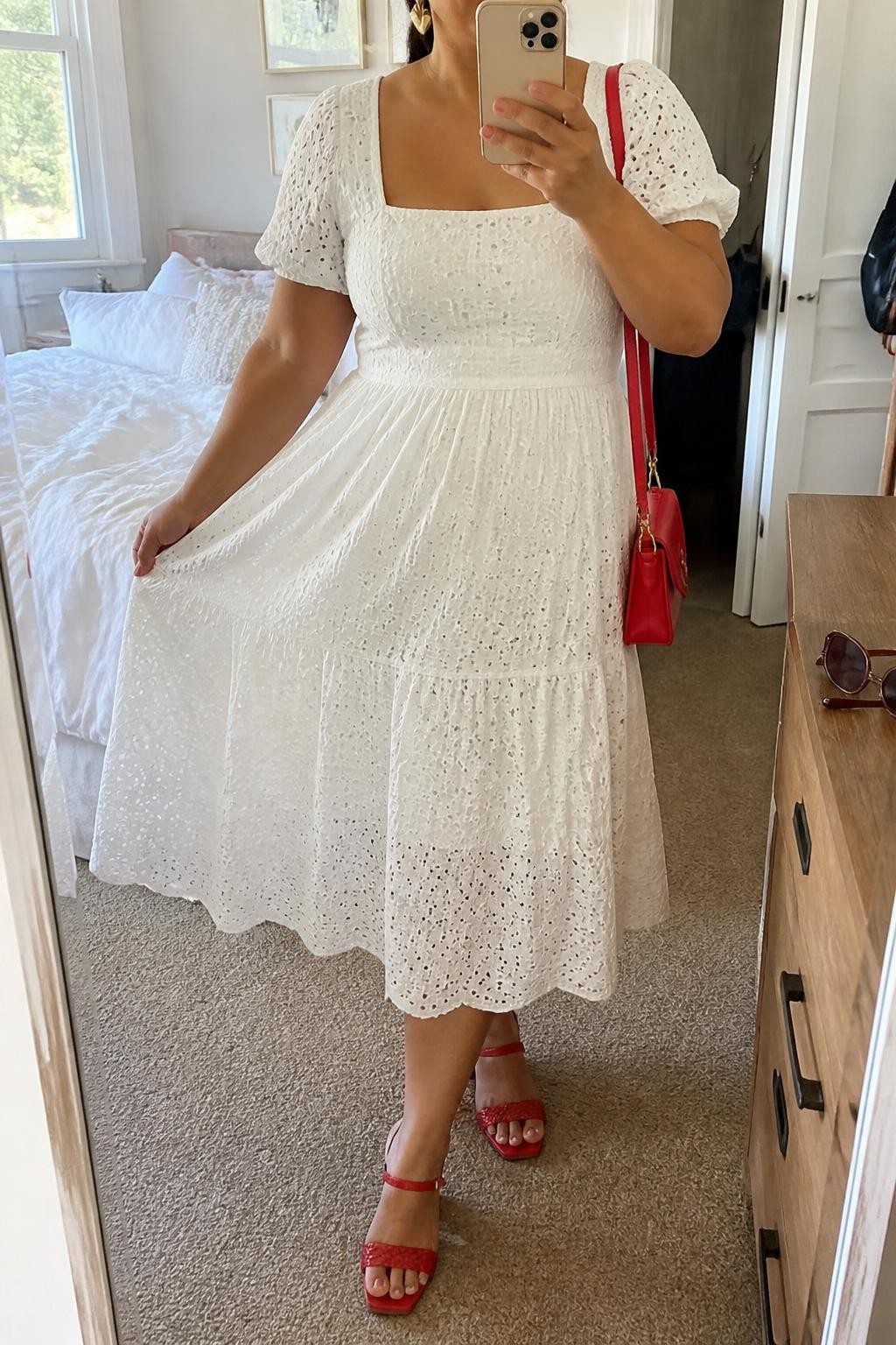 Mirror selfie of a plus size woman wearing a white cotton eyelet midi dress with short puff sleeves, a square neckline, a fitted waist, and a softly flared skirt, red woven block-heel sandals, a small red leather shoulder bag, and gold heart-shaped earrings, clearly showing the exact dress, white eyelet texture, red footwear, and accessories listed, holding a phone in front of a mirror, cropped at the neck so her face does not appear, relaxed playful selfie pose, in a cozy bedroom mirror setup with natural window light, soft pillows on an unmade bed and sunglasses placed on a dresser, realistic mimosa brunch appearance, latest iPhone photo quality.