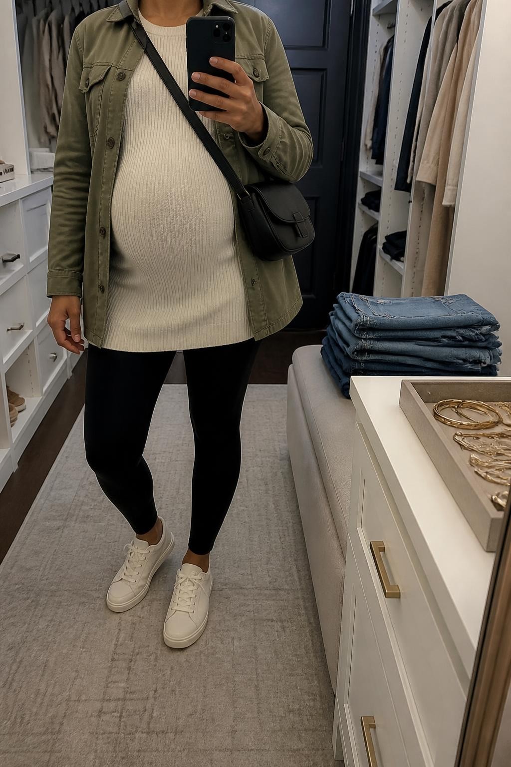 mirror selfie of a Black pregnant woman wearing an open olive cotton utility jacket over a cream longline ribbed maternity sweater, black over-the-bump maternity leggings, cream leather sneakers, and a black nylon crossbody bag adjusted to sit beside the visible baby bump, clearly showing the exact clothing items, colors, fabrics, sneakers, and bag listed in the Outfit Pieces section, holding a phone in front of a mirror, cropped at NECK so the face does not show, relaxed casual maternity selfie pose in a walk-in closet mirror, soft indoor lighting, with folded denim jeans on a bench and a jewelry tray on the vanity, realistic cotton, ribbed knit, stretch fabric, leather, and nylon textures, natural pregnancy posture, latest iPhone photo quality