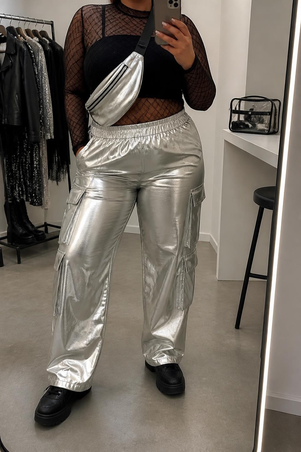 A realistic contemporary fashion mirror selfie featuring a plus-size adult woman in a modern festival outfit. She wears silver metallic cargo pants with flat side pockets, paired with a black long-sleeve mesh fashion top layered over a fully opaque, full-coverage black cropped top. She wears black platform sneakers and a compact silver belt bag worn crossbody. She holds a smartphone in front of a full-length mirror, with the framing cropped below the chin so her face is outside the composition. Relaxed, confident fashion pose in a fashion-studio mirror corner with soft indoor lighting, a clothing rack with garments behind her and a cosmetic bag on the counter. Contemporary festival street style, realistic metallic and mesh textures, detailed cargo pockets, coordinated monochrome accents, natural proportions, authentic indoor lighting, and high-quality modern smartphone photography.