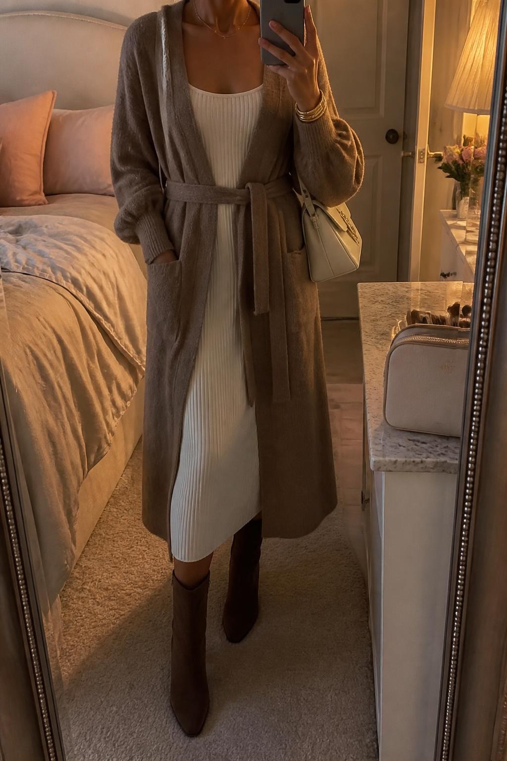 Mirror selfie of a woman wearing a long mocha-brown belted cardigan over a fitted ivory ribbed-knit midi sweater dress, chocolate-brown suede ankle boots, a cream leather shoulder bag, and thin gold bangles, clearly showing the exact clothing items, colors, fabrics, and accessories listed, holding a phone in front of a feminine-themed bedroom mirror, cropped at the neck so the face does not show, relaxed casual selfie pose, warm moody lighting, soft pillows on an unmade bed and a cosmetic bag on the counter, cozy and uncluttered, latest iPhone photo quality.
