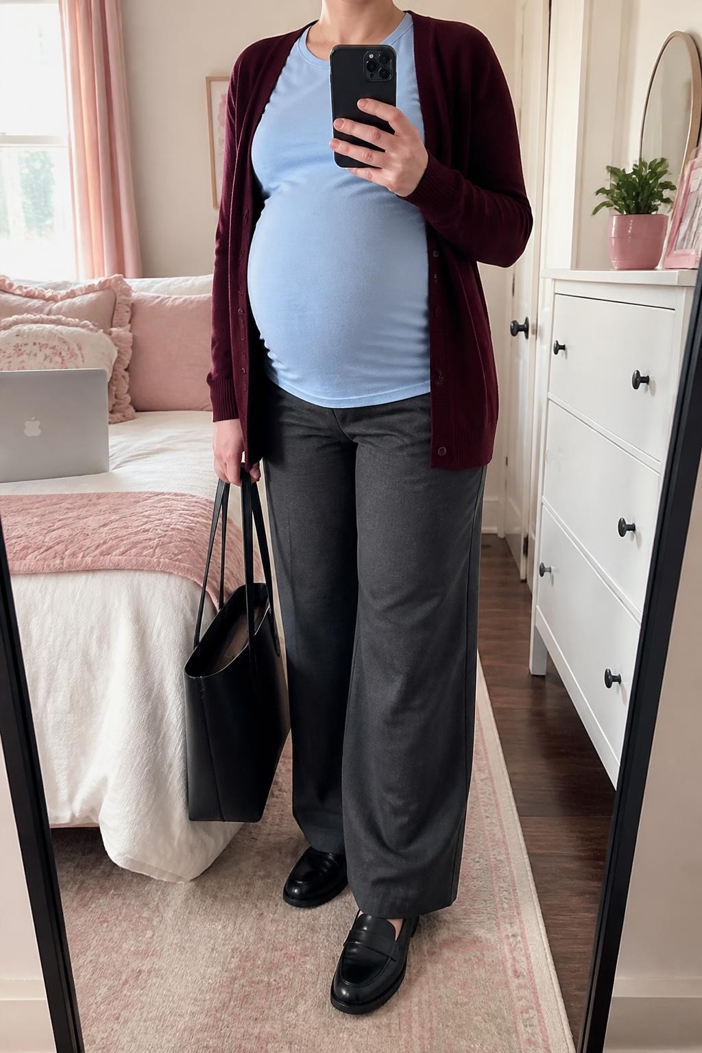 mirror selfie of a pregnant woman wearing an open burgundy soft-knit cardigan over an untucked pale blue stretch-cotton maternity shirt shaped comfortably around her visible baby bump, charcoal wide-leg maternity trousers, black cushioned leather loafers, and a black structured work tote, clearly showing the exact clothing items, colors, fabrics, shoes, and bag listed in the Outfit Pieces section, holding a phone in front of a mirror, cropped at NECK so the face does not show, relaxed professional maternity selfie pose in a feminine-themed bedroom, natural window light, with a laptop on the bed and a small plant on a dresser, realistic knit, stretch cotton, trouser, and leather textures, latest iPhone photo quality