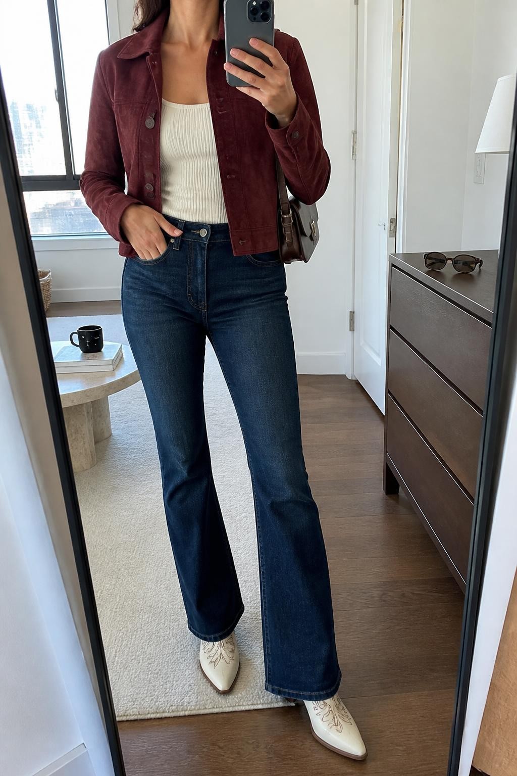 Image Prompt mirror selfie of a woman wearing an open burgundy cropped suede jacket over an ivory fitted ribbed scoop-neck top, dark-wash high-rise flare jeans lightly skimming cream leather cowboy boots with brown stitching while leaving the pointed toes visible, and a chocolate leather shoulder bag, clearly showing the exact clothing items, colors, fabrics, boots, stitching, and bag listed in the Outfit Pieces section, holding a phone in front of a full-length mirror, cropped at NECK so the face does not show, relaxed confident selfie pose in a stylish city apartment, natural window light, with sunglasses placed on a dresser and a coffee mug on a side table, realistic suede, ribbed knit, denim, and leather textures, latest iPhone photo quality