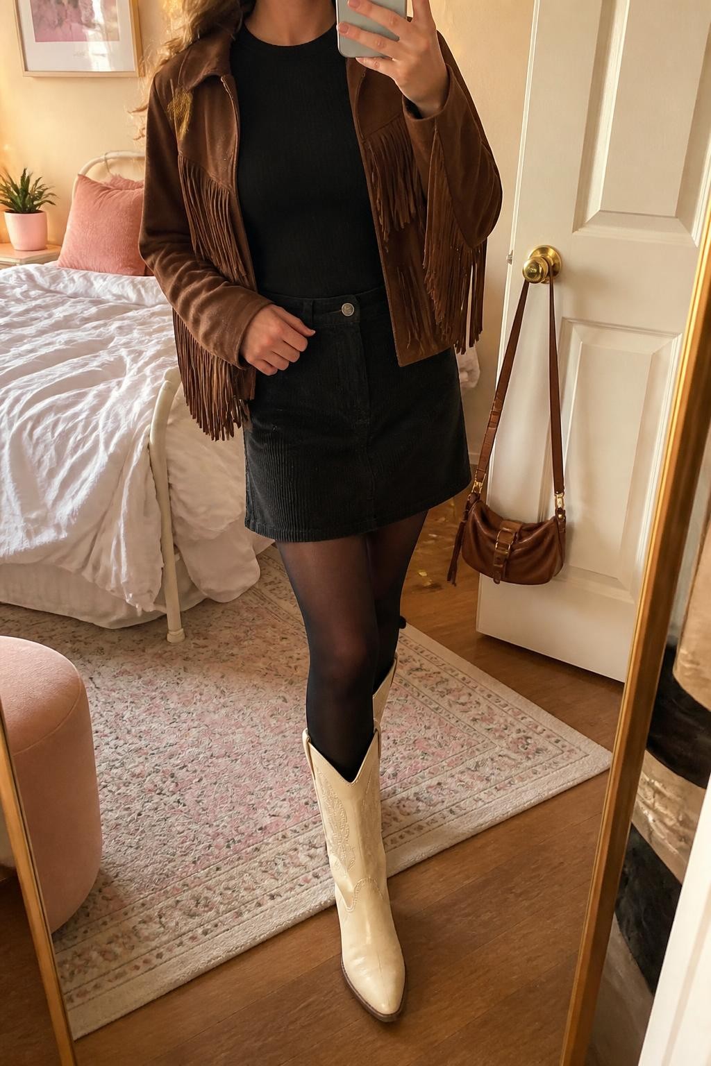 Image Prompt mirror selfie of a woman wearing an open chocolate suede-look fringe jacket over a black fitted ribbed crewneck top, a black A-line corduroy mini skirt, black opaque tights, and cream leather cowboy boots, clearly showing the exact clothing items, colors, fabrics, fringe, tights, and boots listed in the Outfit Pieces section, holding a phone in front of a mirror, cropped at NECK so the face does not show, relaxed playful selfie pose in a feminine-themed bedroom, evening golden hour light, with soft pillows on an unmade bed and a handbag hanging on a door handle, realistic suede-look fringe, ribbed knit, corduroy, tights, and leather textures, latest iPhone photo quality