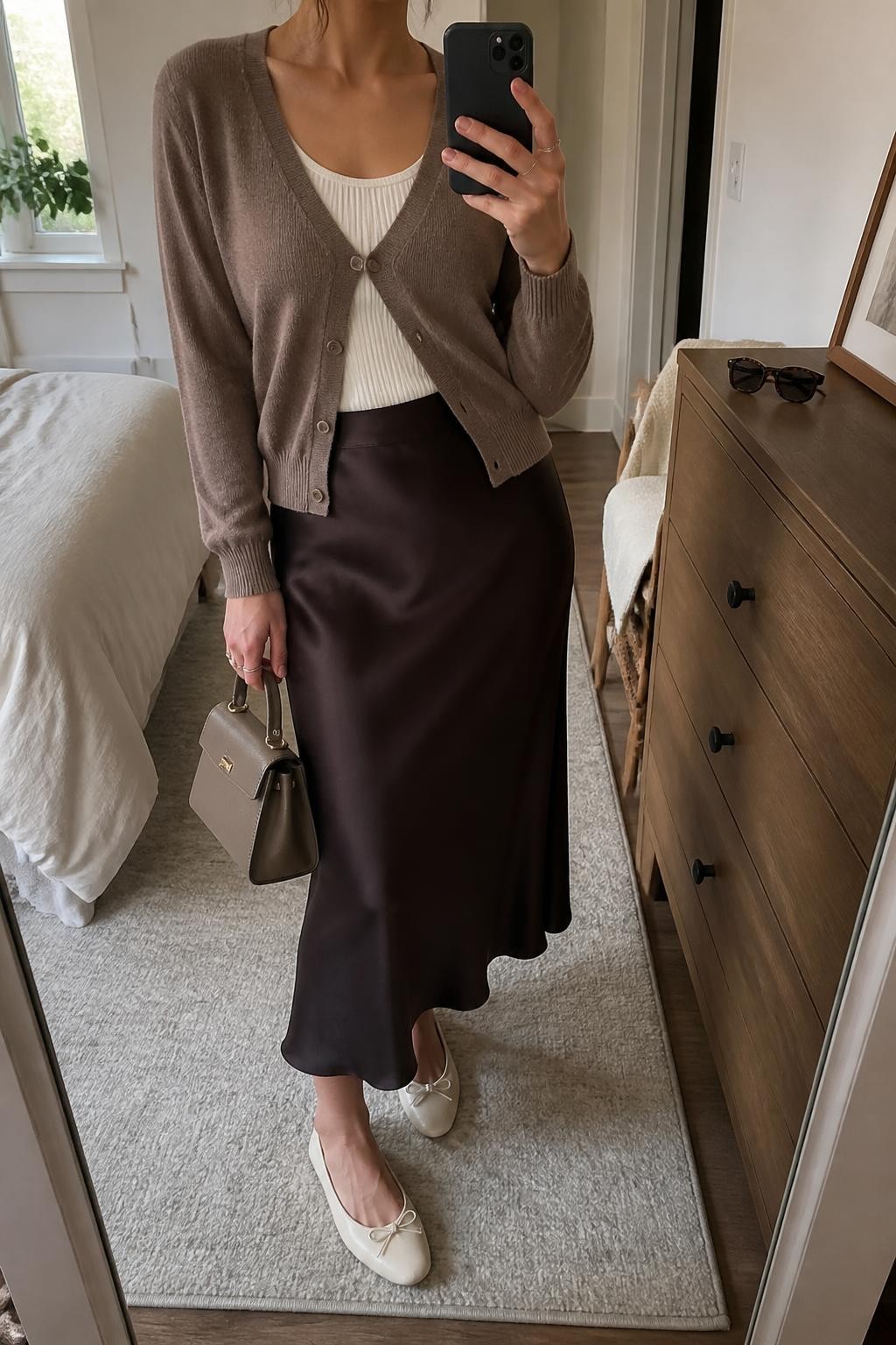 mirror selfie of a woman wearing a mushroom brown fine-knit V-neck cardigan buttoned halfway over an ivory fitted scoop-neck knit top tucked into a dark chocolate satin midi slip skirt, cream leather ballet flats, and a taupe structured mini handbag, clearly showing the exact clothing items, colors, fabrics, shoes, and bag listed in the Outfit Pieces section, holding a phone in front of a mirror, cropped at NECK so the face does not show, relaxed feminine selfie pose in a cozy bedroom mirror setup, natural window light, with a folded blanket on a chair and sunglasses placed on a dresser, realistic fine knit, satin, and leather textures, latest iPhone photo quality