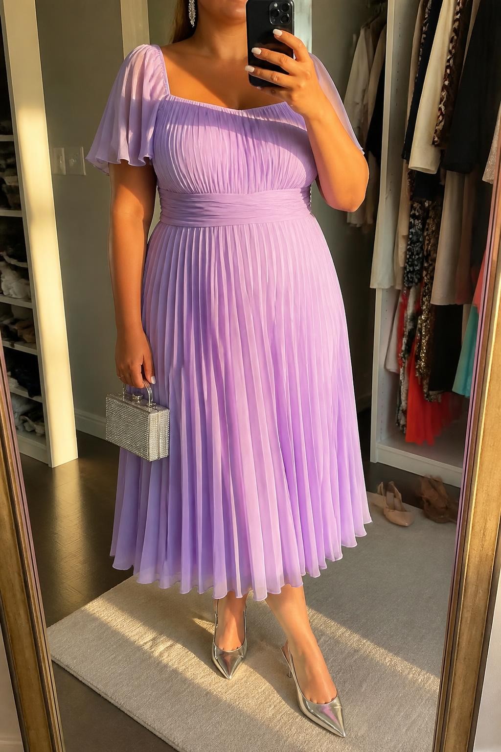 Mirror selfie of a plus size woman wearing a lilac chiffon pleated midi dress with short flutter sleeves, a square neckline, and a softly fitted waist, metallic silver pointed kitten heels, a small silver box handbag, and silver crystal drop earrings, clearly showing the exact dress, lilac chiffon, pleated shape, silver footwear, handbag, and accessories listed, holding a phone in front of a mirror, cropped at the neck so her face does not appear, relaxed polished selfie stance, in a walk-in closet mirror with evening golden-hour lighting, an open closet with clothes visible and a pair of heels on the floor nearby, realistic baby shower celebration outfit, latest iPhone photo quality.