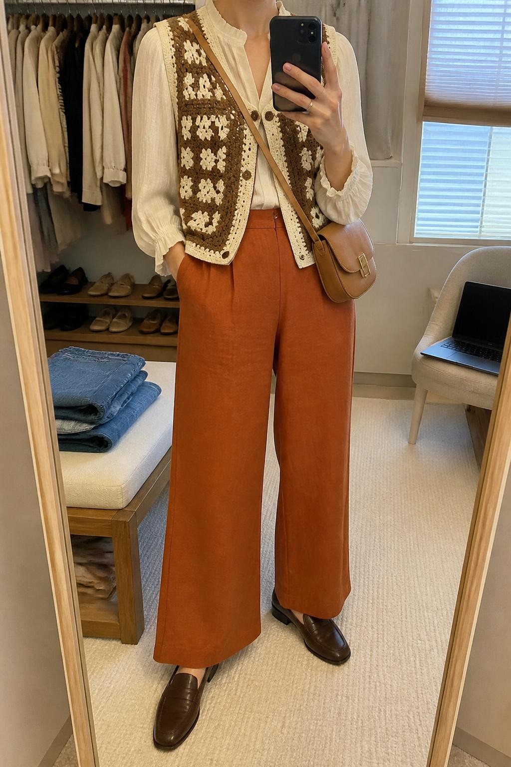 Mirror selfie of a woman wearing rust-orange wide-leg linen-blend pants, a cream long-sleeve blouse, a brown and cream hand-crocheted vest, dark brown leather loafers, and a structured tan crossbody bag, clearly showing the exact clothing items, colors, fabrics, and accessories listed, holding a phone in front of a walk-in closet mirror, cropped at the neck so the face does not show, relaxed casual selfie pose, soft indoor lighting, folded denim jeans on a bench and a laptop on a chair, natural uncluttered atmosphere, latest iPhone photo quality.
