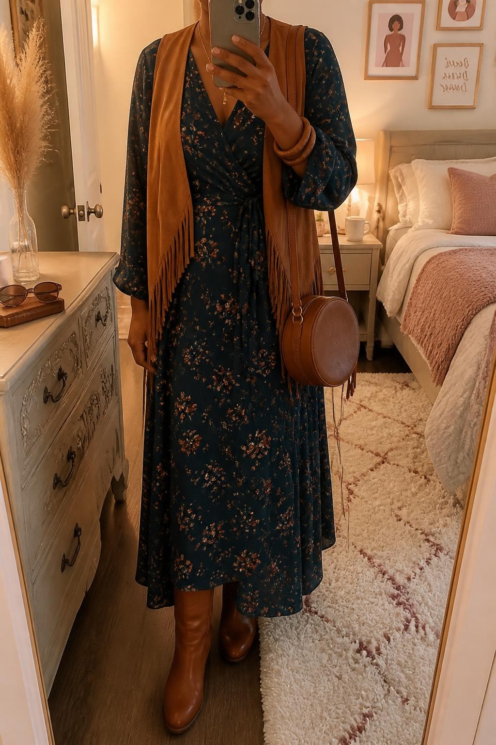 Mirror selfie of a Black woman wearing a dark teal wrap maxi dress with small rust and cream floral prints, a camel suede vest with short fringe, cognac leather knee-high boots, a brown circular crossbody bag, and stacked wooden bangles, clearly showing the exact clothing items, colors, fabrics, and accessories listed, holding a phone in front of a feminine-themed bedroom mirror, cropped at the neck so the face does not show, relaxed casual selfie pose, evening golden-hour lighting, sunglasses placed on a dresser and a coffee mug on a bedside table, warm realistic scene, latest iPhone photo quality.