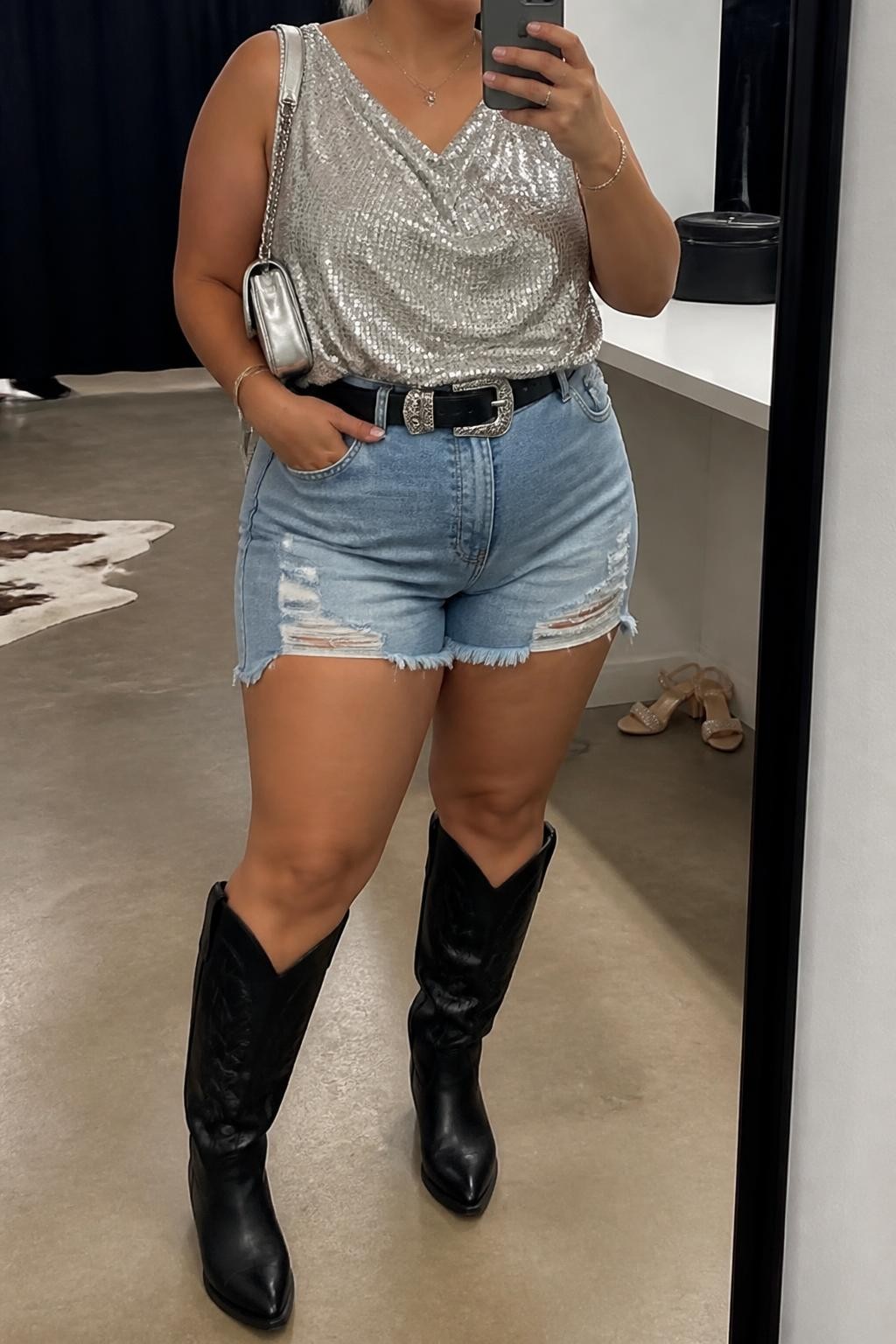 Mirror selfie of a plus size woman wearing a silver sequin sleeveless top with a loose draped fit, high-waisted medium-wash distressed denim shorts with a relaxed mid-thigh length, black leather knee-high cowboy boots, a black Western belt with a silver buckle, and a small silver shoulder bag, clearly showing the exact clothing items, silver sequins, denim distressing, colors, footwear, belt, and accessories listed, holding a phone in front of a mirror, cropped at the neck so the face does not appear, relaxed glamorous selfie stance, in a fashion studio mirror corner with soft indoor lighting, a pair of heels on the floor nearby and a cosmetic bag on the counter, realistic nighttime country concert styling, latest iPhone photo quality.