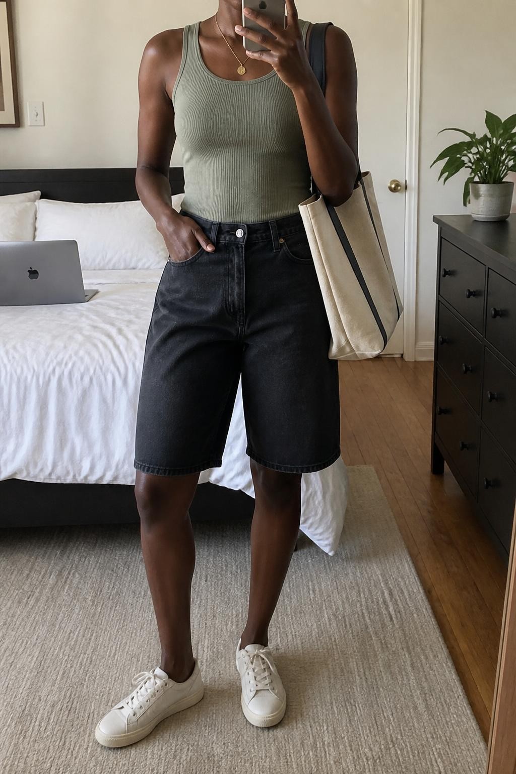 mirror selfie of a woman with deep brown skin wearing clean relaxed knee-length black denim jorts, a fitted sage green ribbed cotton tank top, simple cream low-top sneakers, a natural canvas tote bag with plain black handles and a delicate gold pendant necklace, exactly matching the listed pieces with no additional garments, holding a phone in front of a mirror, cropped at NECK so the face does not show, relaxed casual selfie pose, in a cozy bedroom mirror setup, natural window light, laptop on the bed and a small plant on a dresser, believable casual styling, latest iPhone photo quality.