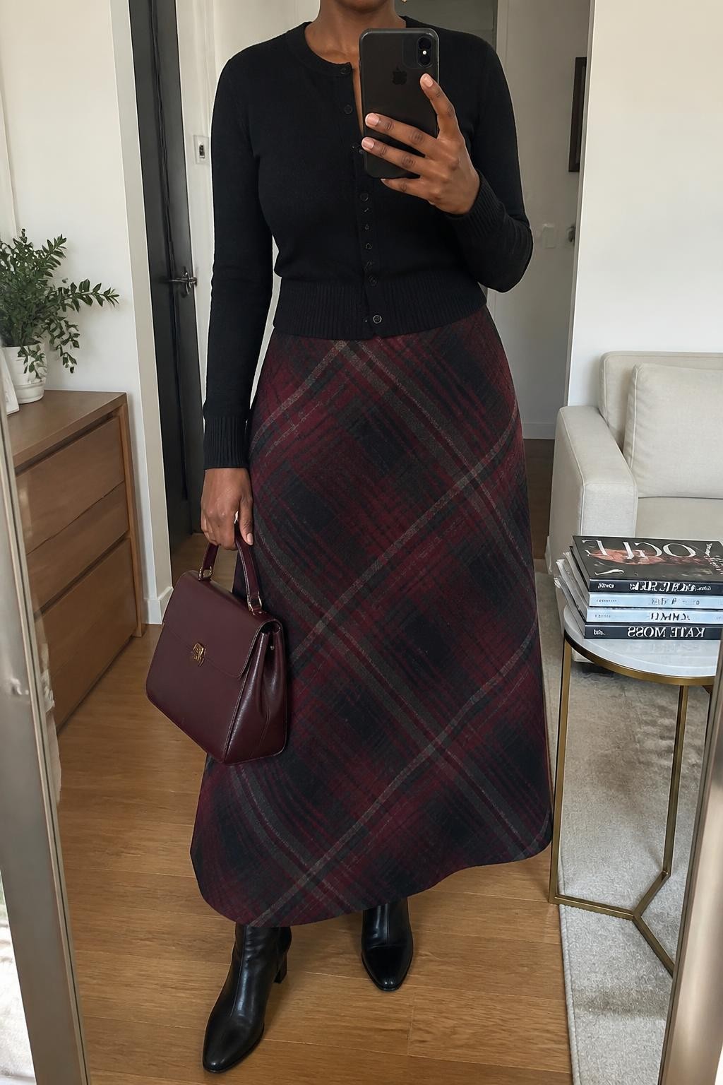 Mirror selfie of a Black woman wearing a long burgundy and charcoal plaid wool skirt, fitted black button-front cardigan, black leather ankle boots, burgundy top-handle bag, and delicate silver stud earrings, clearly showing the exact clothing items, colors, fabrics, and accessories listed, holding a phone in front of a stylish city apartment mirror, cropped at the neck so the face does not show, relaxed casual selfie pose, soft indoor lighting, stack of fashion magazines on a side table and a small plant on a dresser, natural and uncluttered, latest iPhone photo quality.