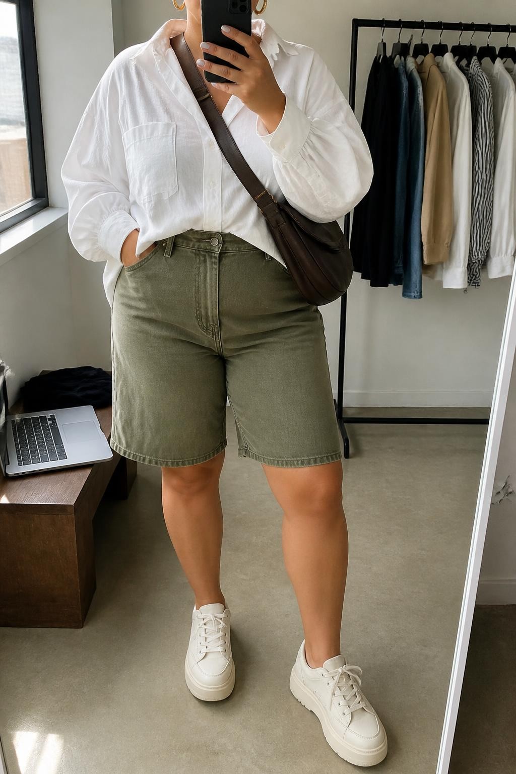 Mirror selfie of a plus size woman wearing olive green high-waisted denim jorts with a relaxed straight cut, an oversized white cotton button-down shirt tucked loosely at the front, cream platform sneakers, a dark brown nylon crossbody bag, and chunky gold hoop earrings, clearly showing the exact clothing items, olive denim, white cotton, colors, footwear, and accessories listed, holding a phone in front of a mirror, cropped at the neck so her face does not show, relaxed casual selfie stance, in a minimal dressing room with natural window light, a laptop on a bench and a clothing rack with garments behind her, realistic weekend outfit, latest iPhone photo quality.