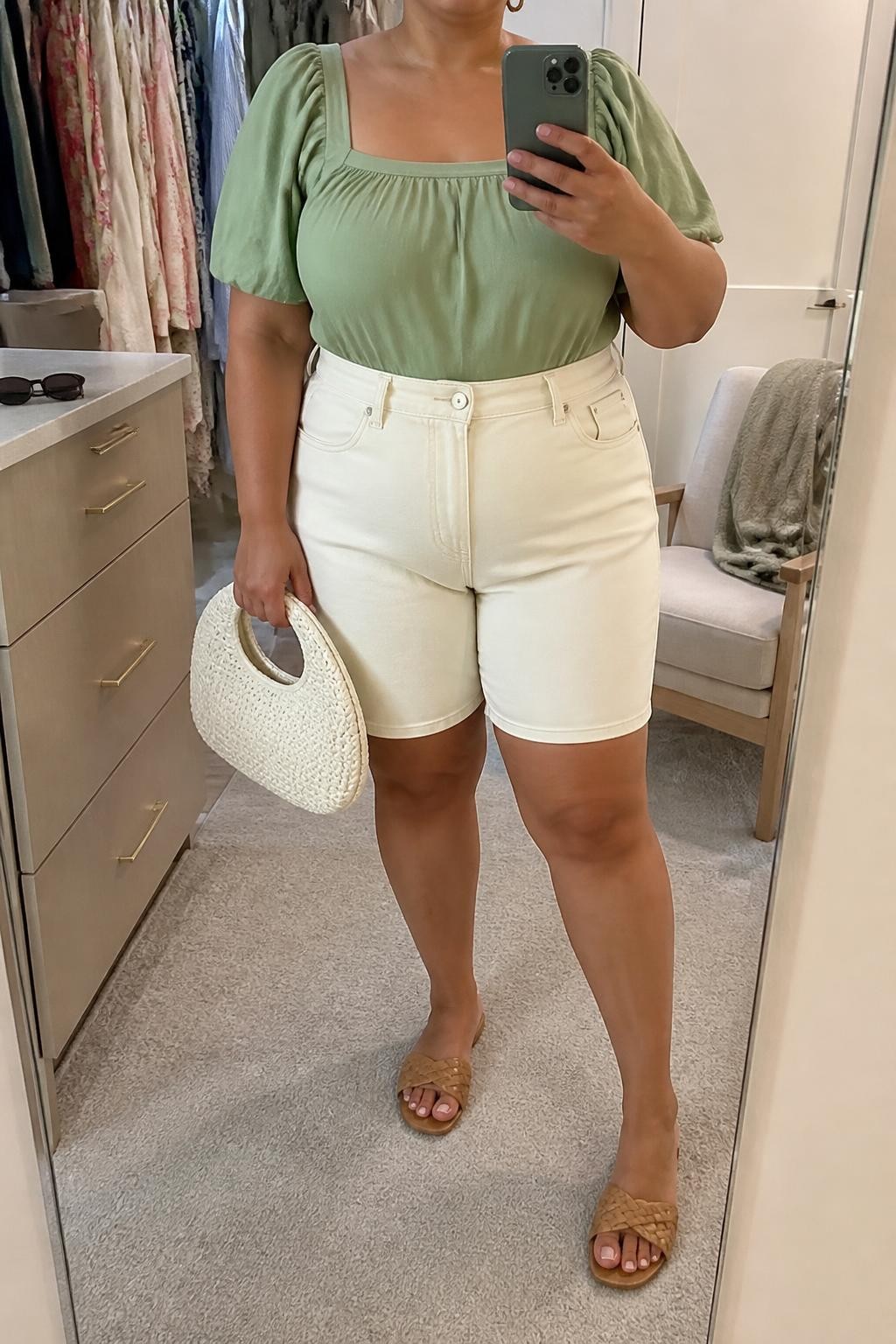 Mirror selfie of a plus size woman wearing ecru high-waisted denim jorts with a clean knee-skimming hem, a sage green cotton blouse with short puff sleeves and a square neckline, tan woven flat sandals, a cream woven handbag, and small gold hoop earrings, clearly showing the exact clothing items, ecru denim, sage fabric, sleeve shape, footwear, and accessories listed, holding a phone in front of a mirror, cropped at the neck so her face does not appear, relaxed feminine selfie stance, in a walk-in closet mirror with soft indoor lighting, sunglasses placed on a dresser and a folded blanket on a chair, realistic brunch outfit, latest iPhone photo quality.