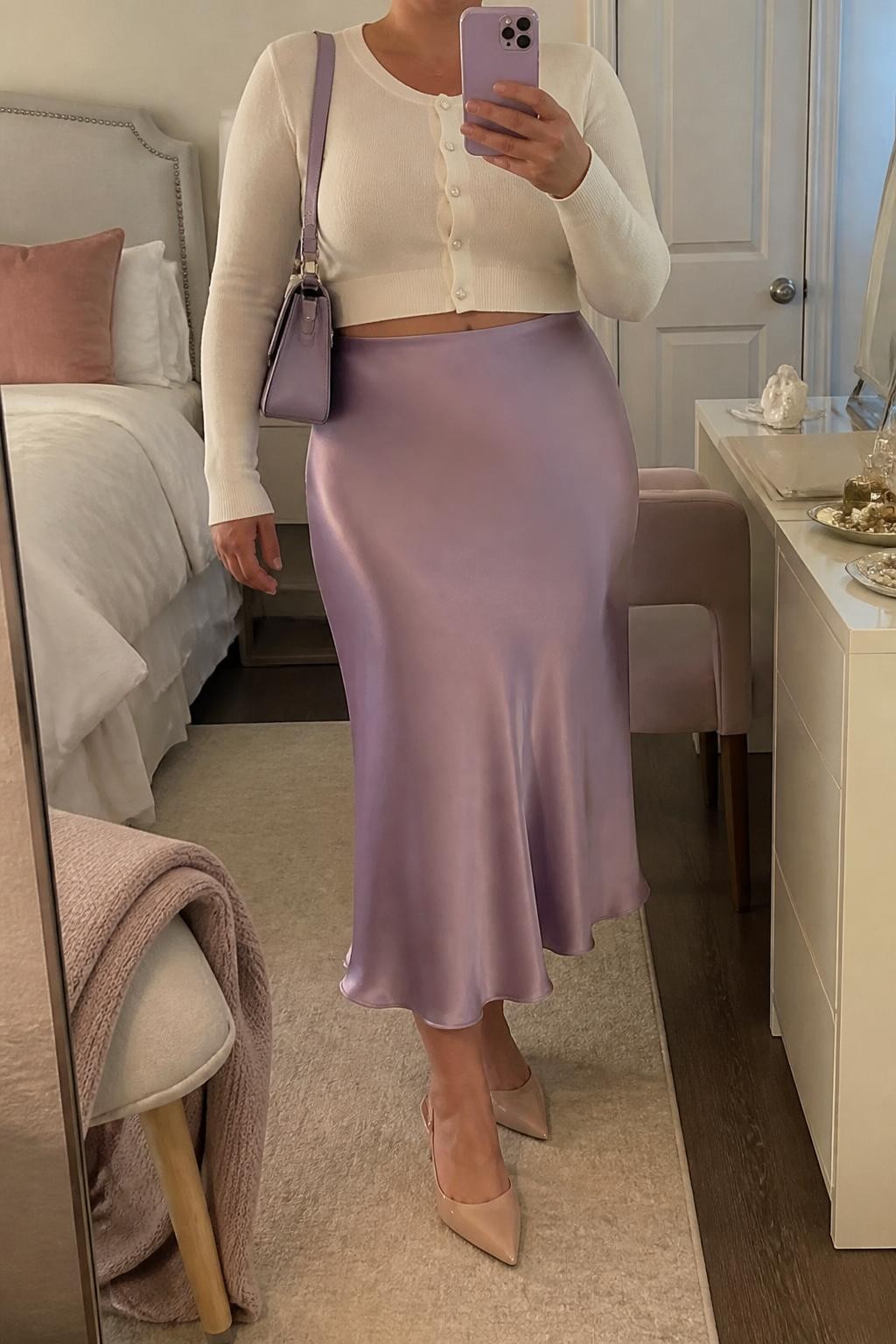 Mirror selfie of a plus size woman wearing a lavender high-waisted satin midi skirt with a fluid bias-cut shape, a fitted ivory cropped cardigan buttoned at the front, nude pointed kitten heels, a structured lavender shoulder bag, and pearl stud earrings, clearly showing the exact clothing items, lavender satin, ivory knit, nude footwear, and accessories listed, holding a phone in front of a mirror, cropped at the neck so her face does not show, relaxed elegant selfie stance, in a cozy bedroom mirror setup with soft indoor lighting, a folded blanket on a chair and a jewelry tray on the vanity, realistic baby shower guest outfit, latest iPhone photo quality.