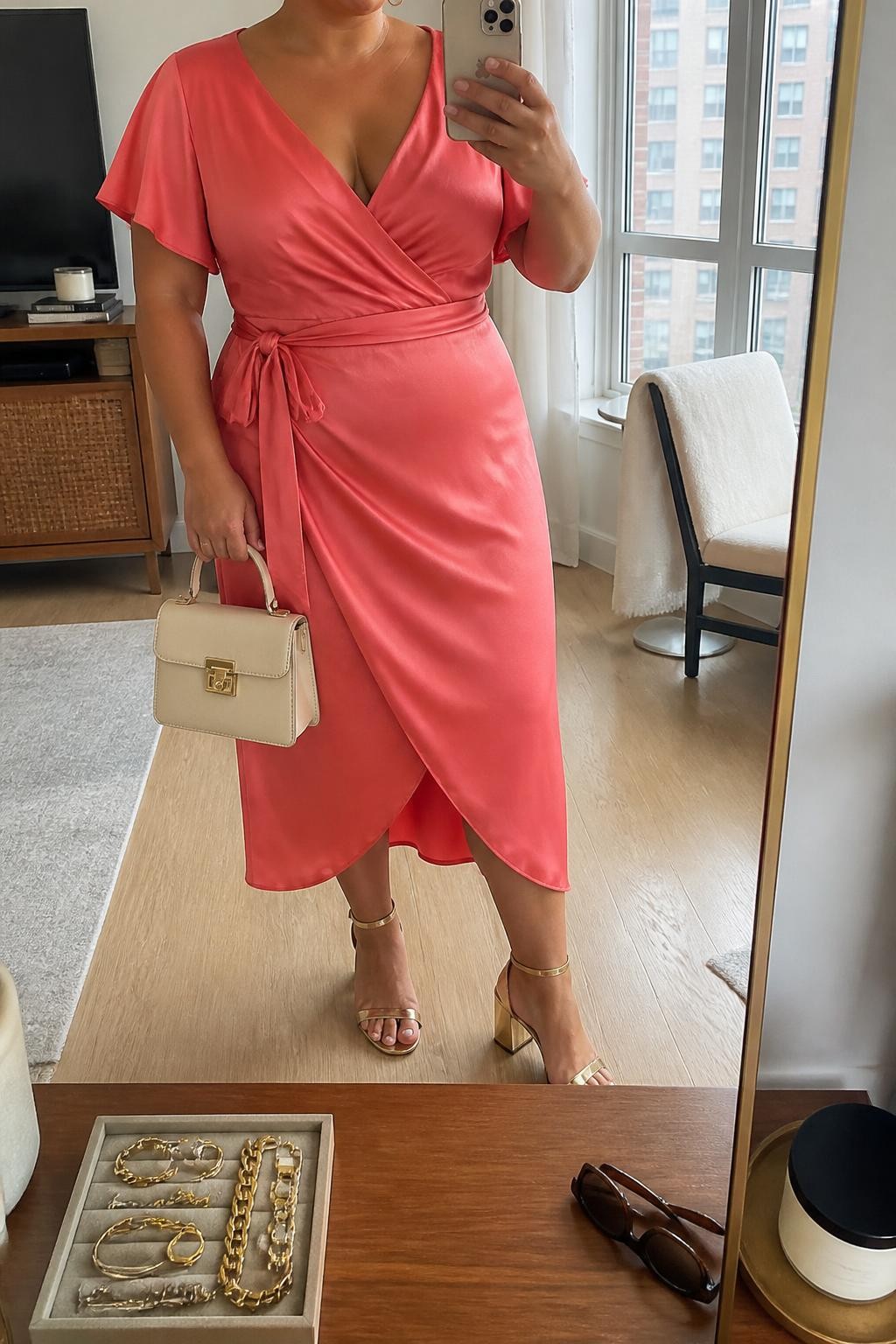 Mirror selfie of a plus size woman wearing a coral pink satin wrap midi dress with short flutter sleeves, a softly draped V-neckline, and a matching waist tie, metallic gold block-heel sandals, a cream structured mini handbag, and delicate gold hoop earrings, clearly showing the exact clothing items, coral satin, wrap shape, colors, footwear, and accessories listed, holding a phone in front of a mirror, cropped at the neck so the face does not show, relaxed feminine selfie pose, in a stylish city apartment with natural window light, a jewelry tray on the dresser and sunglasses placed nearby, natural unstaged brunch appearance, latest iPhone photo quality.
