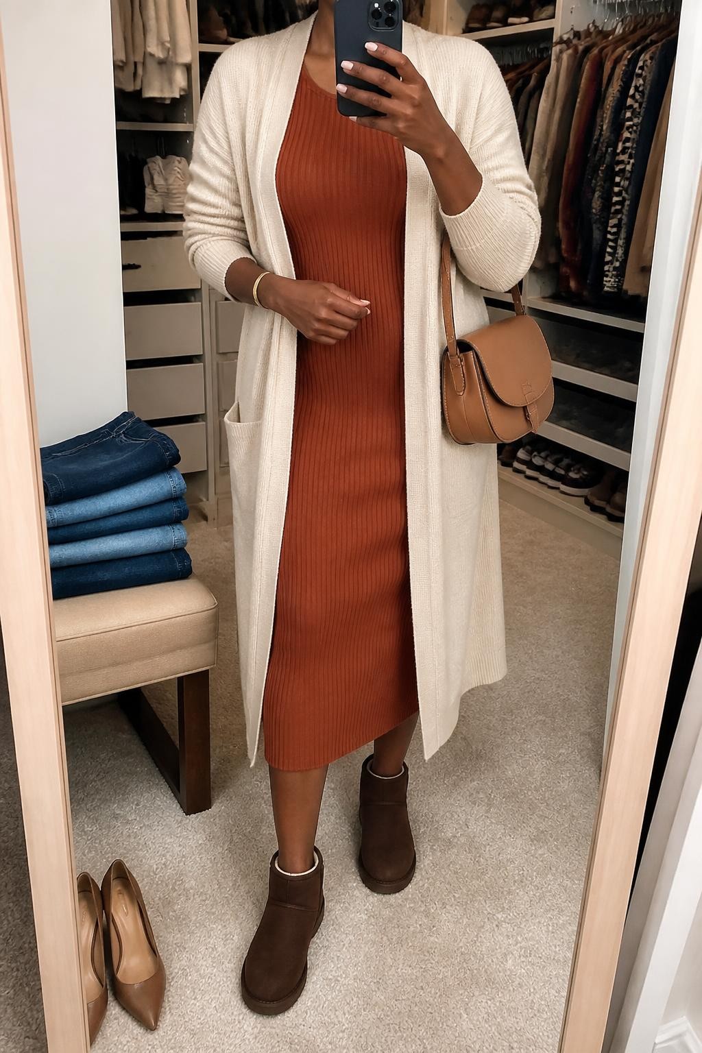 mirror selfie of a Black woman wearing a rust ribbed long-sleeve midi dress with a small space between the hem and chocolate brown mini Ugg boots, a fully open cream longline cardigan, a tan leather saddle bag, and a slim gold cuff bracelet, clearly showing the exact clothing items, colors, fabrics, boots, bag, and bracelet listed in the Outfit Pieces section, holding a phone in front of a mirror, cropped at NECK so the face does not show, relaxed polished selfie pose in a walk-in closet mirror, soft indoor lighting, with folded denim jeans on a bench and a pair of heels on the floor nearby, realistic ribbed knit, soft cardigan knit, suede, and leather textures, latest iPhone photo quality