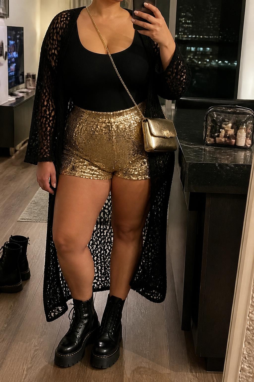 Mirror selfie of a plus size woman wearing metallic gold high-waisted sequin shorts with relaxed thigh openings, a fitted black scoop-neck bodysuit, a long black crochet duster worn open, black platform combat boots, and a small gold crossbody bag, clearly showing the exact clothing items, gold sequins, black crochet, fitted bodysuit, footwear, and accessories listed, holding a phone in front of a mirror, cropped at the neck so the face does not show, relaxed glamorous selfie pose, in a stylish city apartment with warm moody lighting, a cosmetic bag on the counter and a pair of boots on the floor nearby, realistic evening festival outfit, latest iPhone photo quality.
