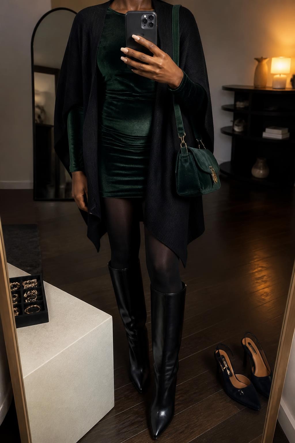 mirror selfie of a Black woman wearing an open black soft-knit cape cardigan over a forest green velvet long-sleeve mini dress, black opaque tights, black leather knee-high boots, and a forest green velvet shoulder bag, clearly showing the exact clothing items, colors, fabrics, tights, boots, and bag listed in the Outfit Pieces section, holding a phone in front of a mirror, cropped at NECK so the face does not show, relaxed confident selfie pose in a fashion studio mirror corner, warm moody lighting, with a jewelry tray on a nearby surface and a pair of heels on the floor nearby, realistic soft knit, velvet, tights, and leather textures, latest iPhone photo quality
