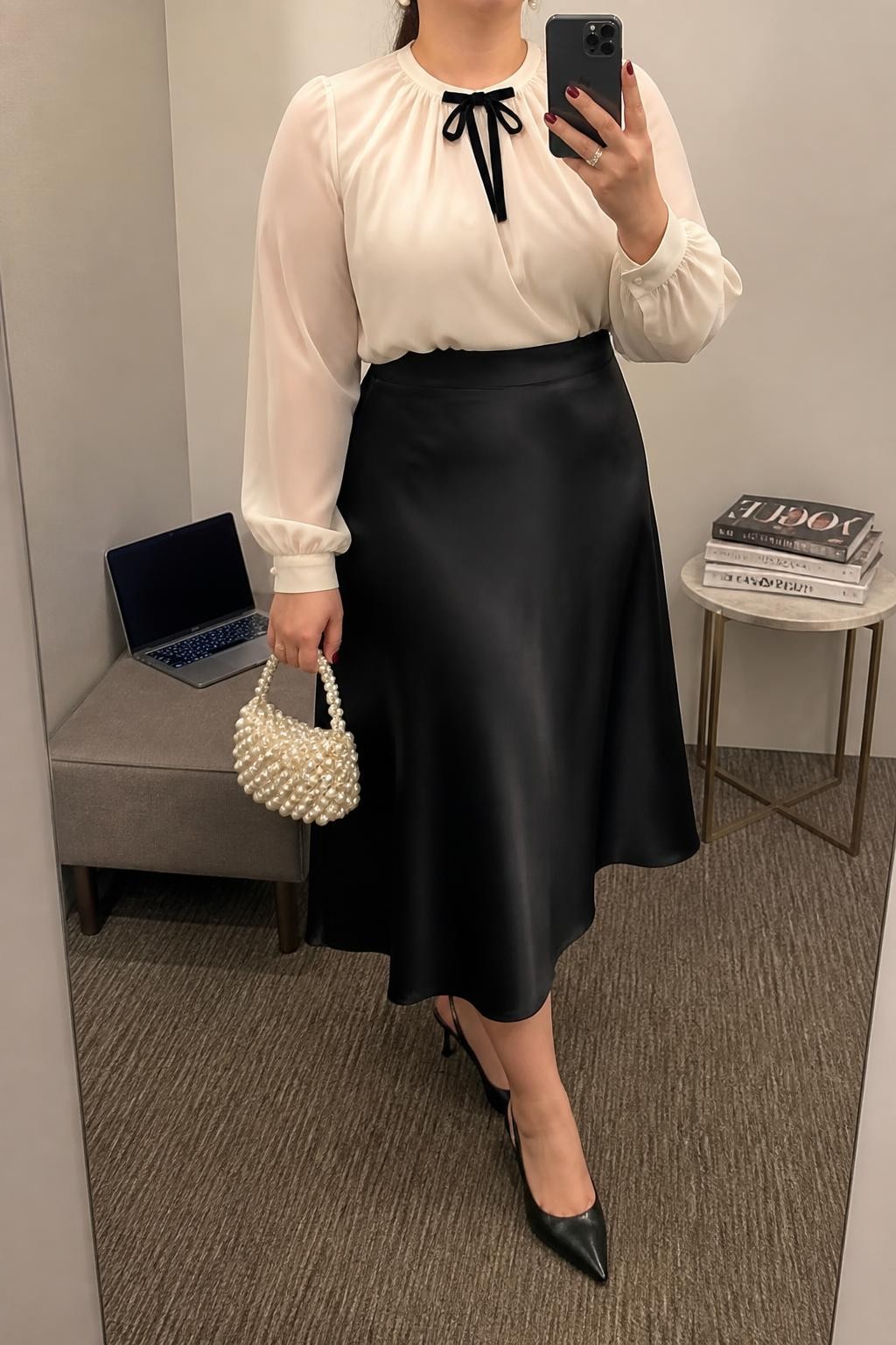 Mirror selfie of a plus size woman wearing a black high-waisted satin A-line midi skirt, an ivory chiffon blouse with long sleeves and a small black velvet bow at the neckline, black pointed slingback heels, a pearl-covered mini handbag, and pearl stud earrings, clearly showing the exact clothing items, black satin, ivory chiffon, bow detail, footwear, and accessories listed, holding a phone in front of a mirror, cropped at the neck so her face does not show, relaxed sophisticated selfie stance, in a minimal dressing room with soft indoor lighting, a laptop on a bench and a stack of fashion magazines on a side table, realistic office event outfit, latest iPhone photo quality.