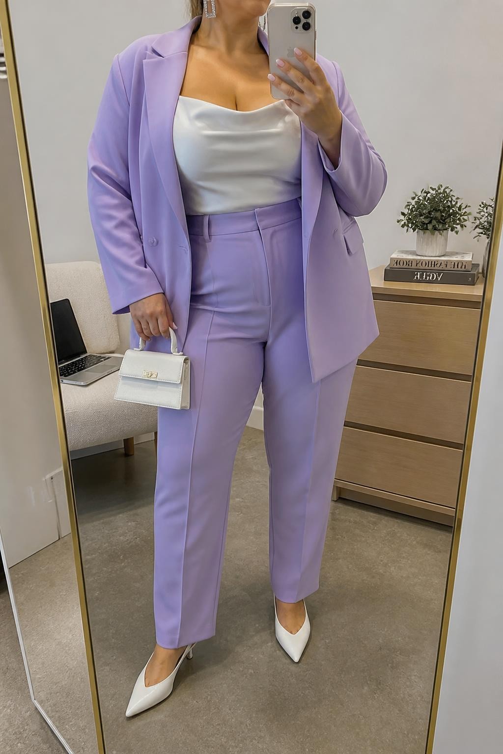 Mirror selfie of a plus size woman wearing a lavender tailored blazer with matching high-waisted straight-leg trousers, a fitted white satin camisole with wide supportive straps, white pointed kitten heels, a structured white mini handbag, and silver rectangular earrings, clearly showing the exact suit, lavender color, white satin, footwear, and accessories listed, holding a phone in front of a mirror, cropped at the neck so the face does not show, relaxed polished selfie pose, in a fashion studio mirror corner with soft indoor lighting, a laptop on a bench and a small plant on a dresser, realistic upscale brunch outfit, latest iPhone photo quality.