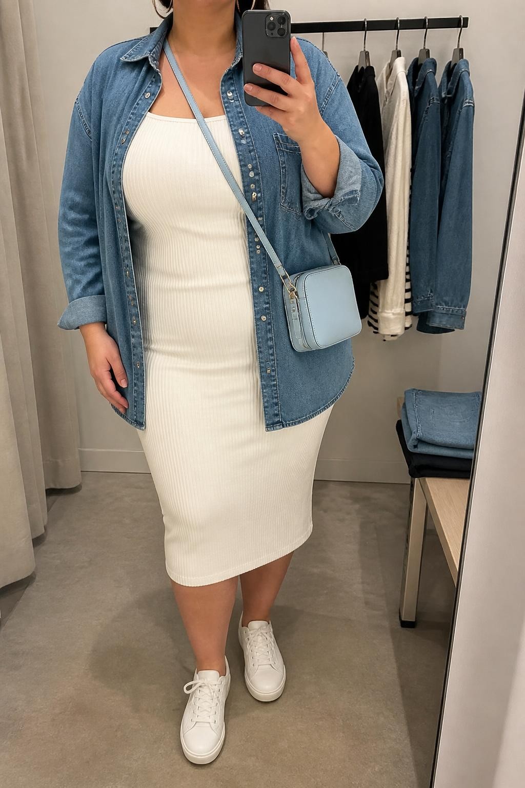Mirror selfie of a plus size woman wearing an oversized medium-wash denim button-down shirt worn open over a fitted white ribbed midi dress with short sleeves and a square neckline, white leather sneakers, a small light blue crossbody bag, and silver hoop earrings, clearly showing the exact clothing layers, denim wash, white ribbed dress, footwear, and accessories listed, holding a phone in front of a mirror, cropped at the neck so the face does not appear, relaxed casual selfie pose, in a minimal dressing room with soft indoor lighting, a clothing rack with garments behind her and folded denim jeans on a bench, realistic daytime outfit, latest iPhone photo quality.