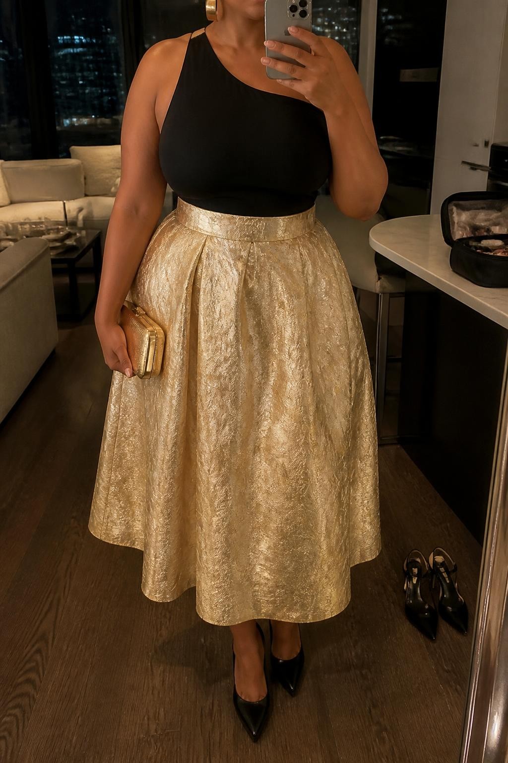 Mirror selfie of a plus size woman wearing a metallic gold high-waisted jacquard midi skirt with a softly structured A-line shape, a fitted black one-shoulder top with a secure wide strap, black pointed platform heels, a small gold hard-case clutch, and chunky gold cuff earrings, clearly showing the exact clothing items, gold jacquard texture, black asymmetric top, footwear, and accessories listed, holding a phone in front of a mirror, cropped at the neck so her face is hidden, relaxed dramatic selfie stance, in a stylish city apartment with warm moody lighting, a pair of heels on the floor nearby and a cosmetic bag on the counter, realistic formal celebration outfit, latest iPhone photo quality.