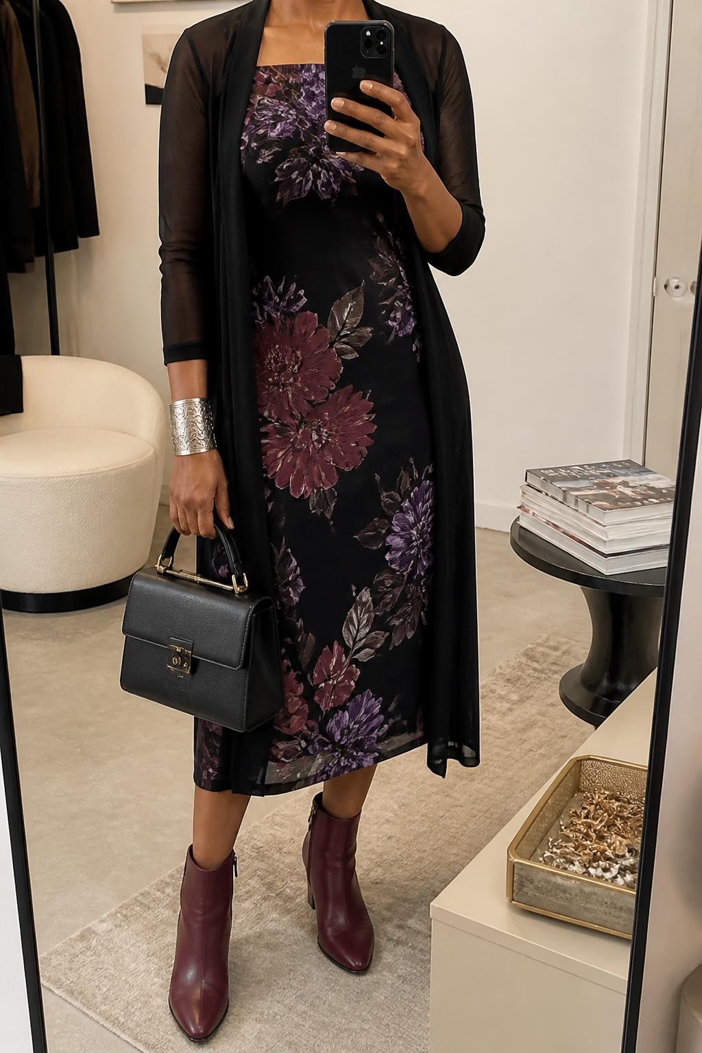 mirror selfie of a Black woman wearing a black midi dress with a clear burgundy and purple floral print, an open black sheer mesh longline cardigan, burgundy leather ankle boots, a black structured top-handle bag, and an antique silver cuff bracelet, clearly showing the exact clothing items, colors, floral print, fabrics, boots, bag, and bracelet listed in the Outfit Pieces section, holding a phone in front of a mirror, cropped at NECK so the face does not show, relaxed feminine selfie pose in a fashion studio mirror corner, soft indoor lighting, with a jewelry tray on a nearby surface and a stack of fashion magazines on a side table, realistic dress fabric, sheer mesh, leather, and metal textures, latest iPhone photo quality