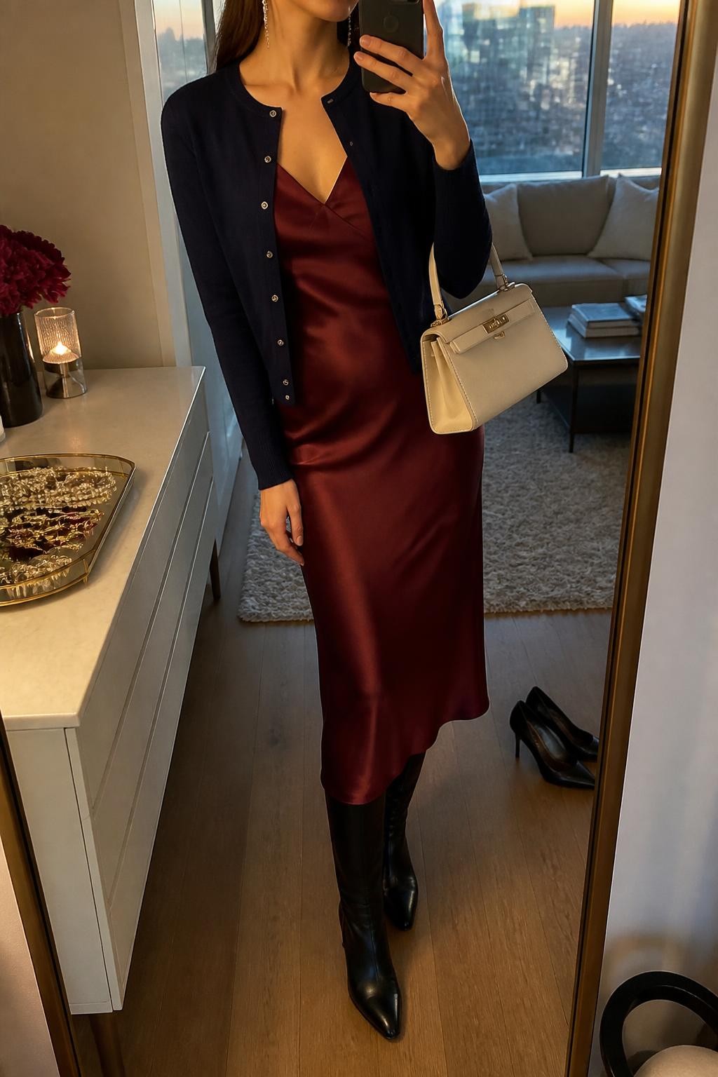 Mirror selfie of a woman wearing a fitted navy cardigan layered over a burgundy satin midi slip dress, black leather knee-high boots, a structured cream handbag, and delicate silver drop earrings, clearly showing the exact clothing items, colors, fabrics, and accessories listed, holding a phone in front of a stylish city apartment mirror, cropped at the neck so the face does not show, relaxed casual selfie pose, evening golden-hour lighting, a jewelry tray on the vanity and a pair of heels on the floor nearby, elegant natural atmosphere, latest iPhone photo quality.