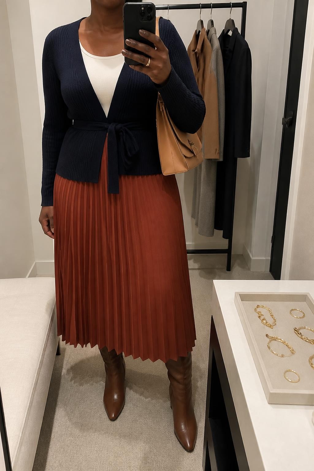 mirror selfie of a Black curvy woman wearing a navy fine-knit belted cardigan tied slightly above the waist over an ivory fitted scoop-neck top, a rust pleated midi skirt, brown leather knee-high boots, and a tan structured shoulder bag, clearly showing the exact clothing items, colors, fabrics, boots, and bag listed in the Outfit Pieces section, holding a phone in front of a mirror, cropped at NECK so the face does not show, relaxed confident selfie pose in a minimal dressing room, soft indoor lighting, with a jewelry tray on the vanity and a clothing rack with garments behind her, realistic knit, pleated fabric, and leather textures, latest iPhone photo quality