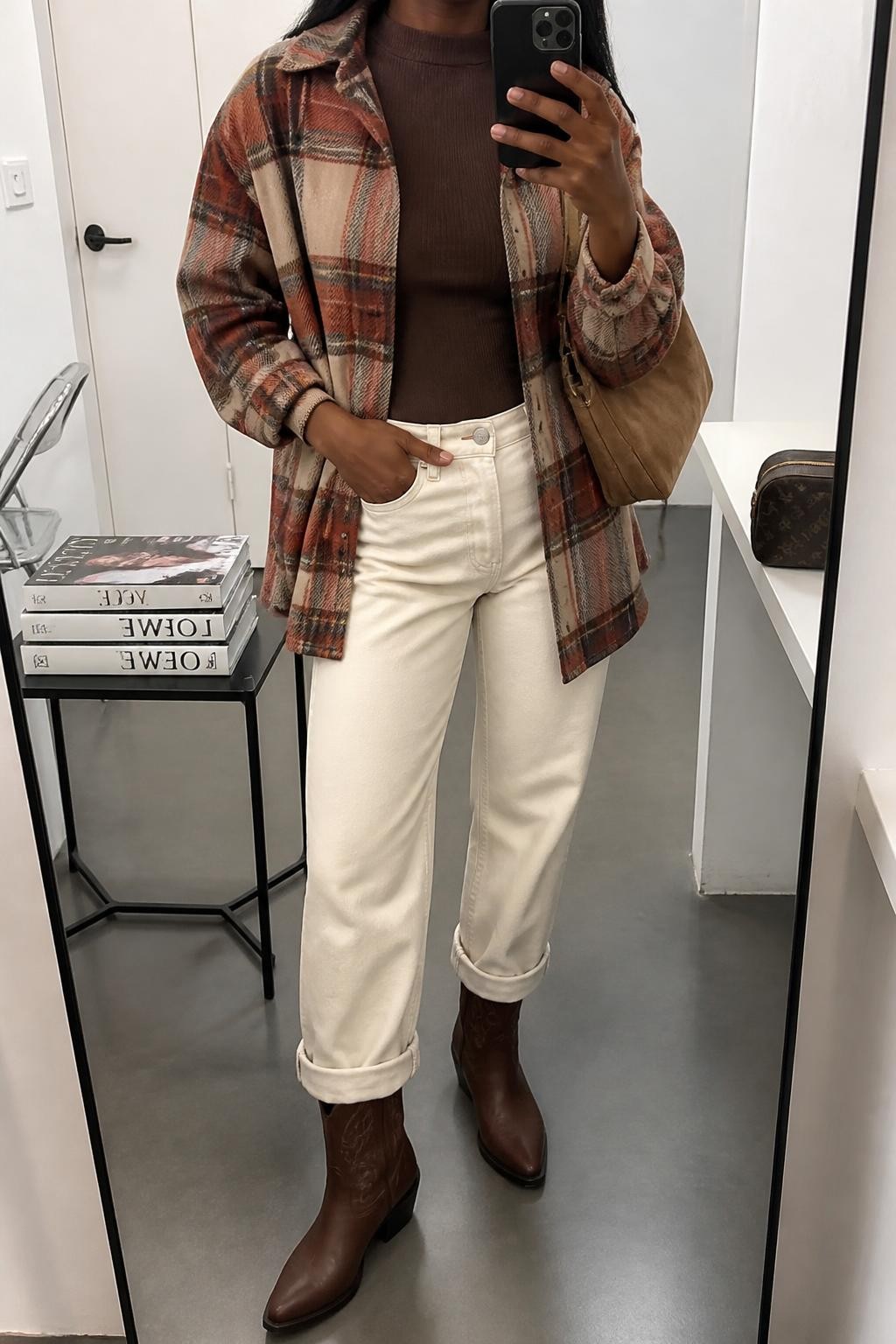 mirror selfie of a Black woman wearing an open brown and rust plaid wool-blend shacket over a chocolate fitted ribbed mock-neck top, ecru straight-leg jeans cuffed once above chestnut leather cowboy boots, and a tan suede shoulder bag, clearly showing the exact clothing items, colors, plaid pattern, fabrics, boots, and bag listed in the Outfit Pieces section, holding a phone in front of a mirror, cropped at NECK so the face does not show, relaxed casual selfie pose in a fashion studio mirror corner, soft indoor lighting, with a stack of fashion magazines on a side table and a cosmetic bag on a counter, realistic wool, ribbed knit, denim, leather, and suede textures, latest iPhone photo quality
