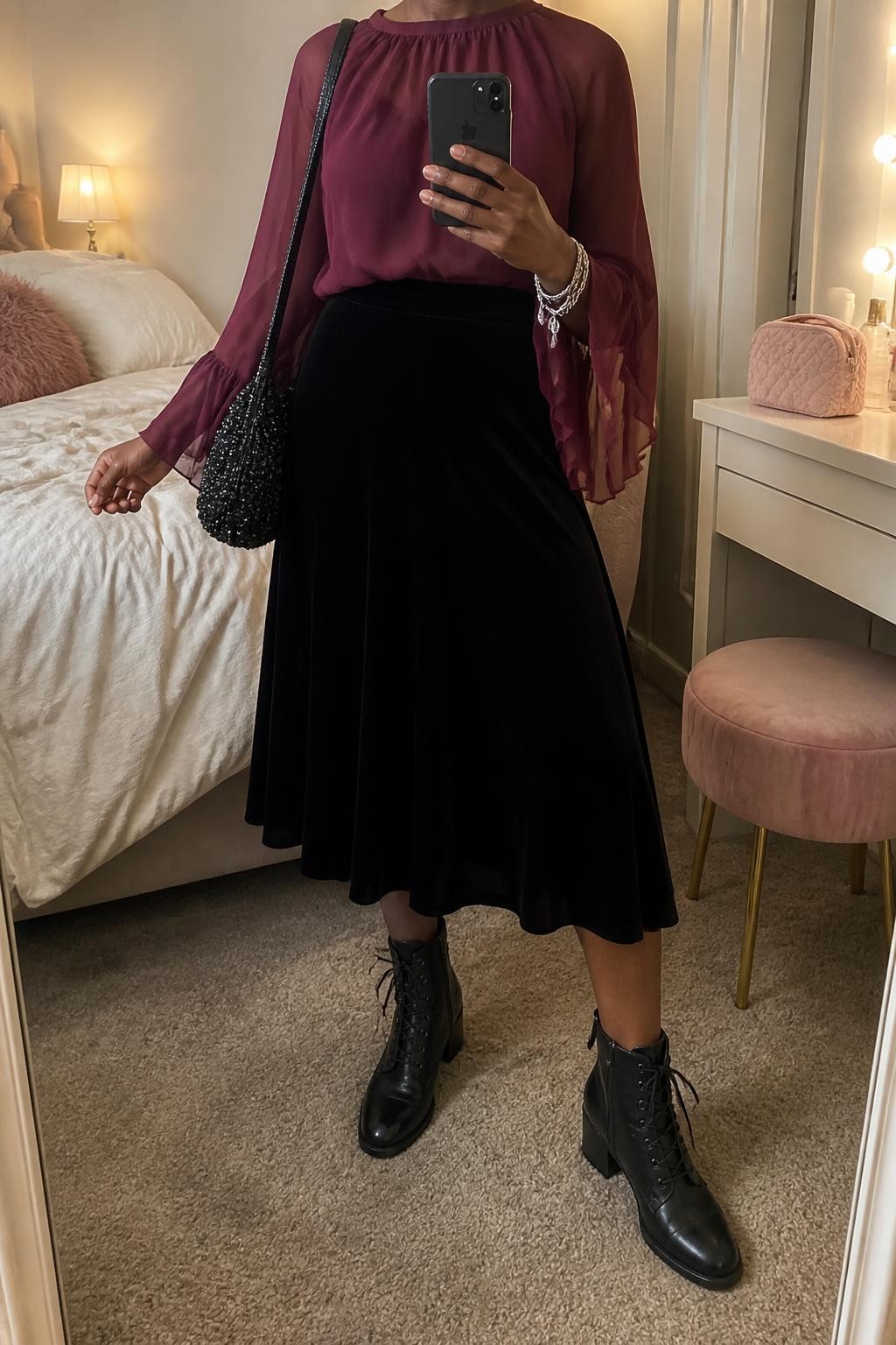 mirror selfie of a Black woman wearing a burgundy chiffon blouse with flowing bell sleeves tucked into a black A-line velvet midi skirt, black leather lace-up ankle boots, a black beaded shoulder bag, and layered silver chain bracelets, clearly showing the exact clothing items, colors, fabrics, boots, bag, and bracelets listed in the Outfit Pieces section, holding a phone in front of a mirror, cropped at NECK so the face does not show, relaxed polished selfie pose in a feminine-themed bedroom, warm moody lighting, with soft pillows on an unmade bed and a cosmetic bag on the vanity, realistic chiffon, velvet, leather, beadwork, and silver textures, latest iPhone photo quality